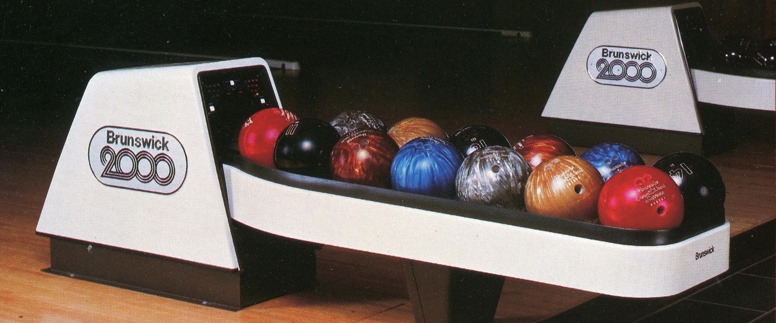 Bowling ball return system labeled "Brunswick 2000" on a wooden floor, holding an array of colorful bowling balls including red, blue, black, and gold.