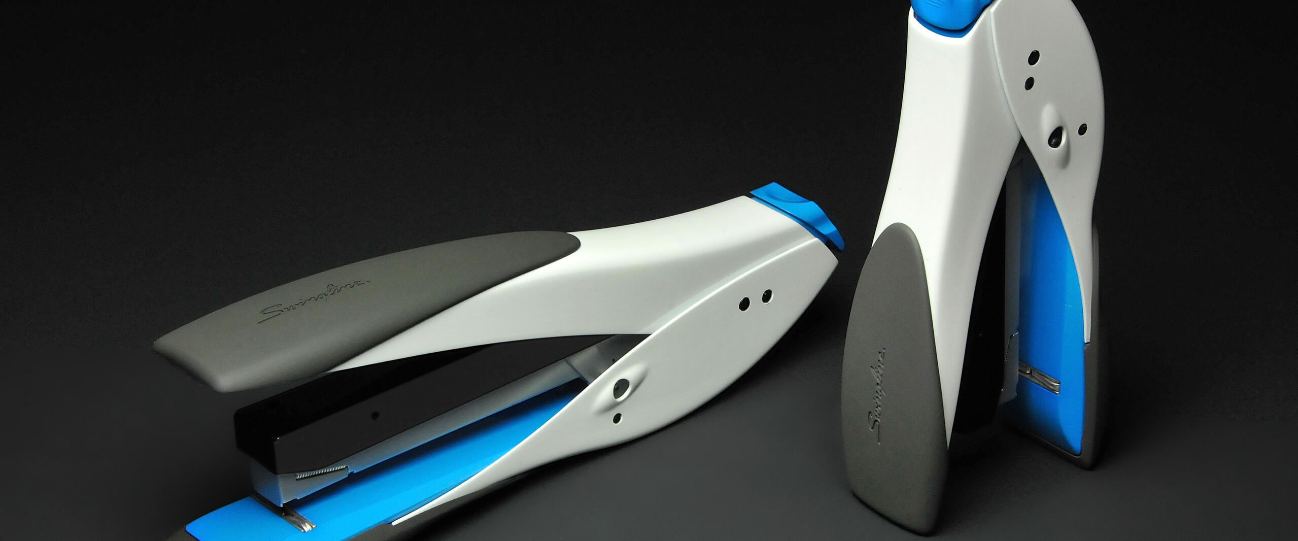 Two modern staplers in white, gray, and blue on a black background, one lying down and one standing.