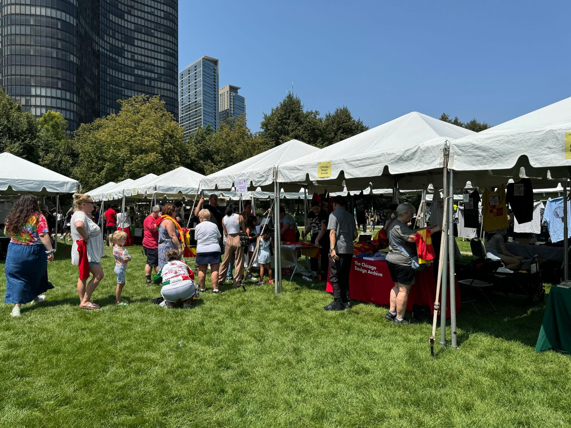 Outdoor event with people browsing stalls under white tents on a grass field. Urban skyscrapers and trees form the background. A sunny day with a clear blue sky.