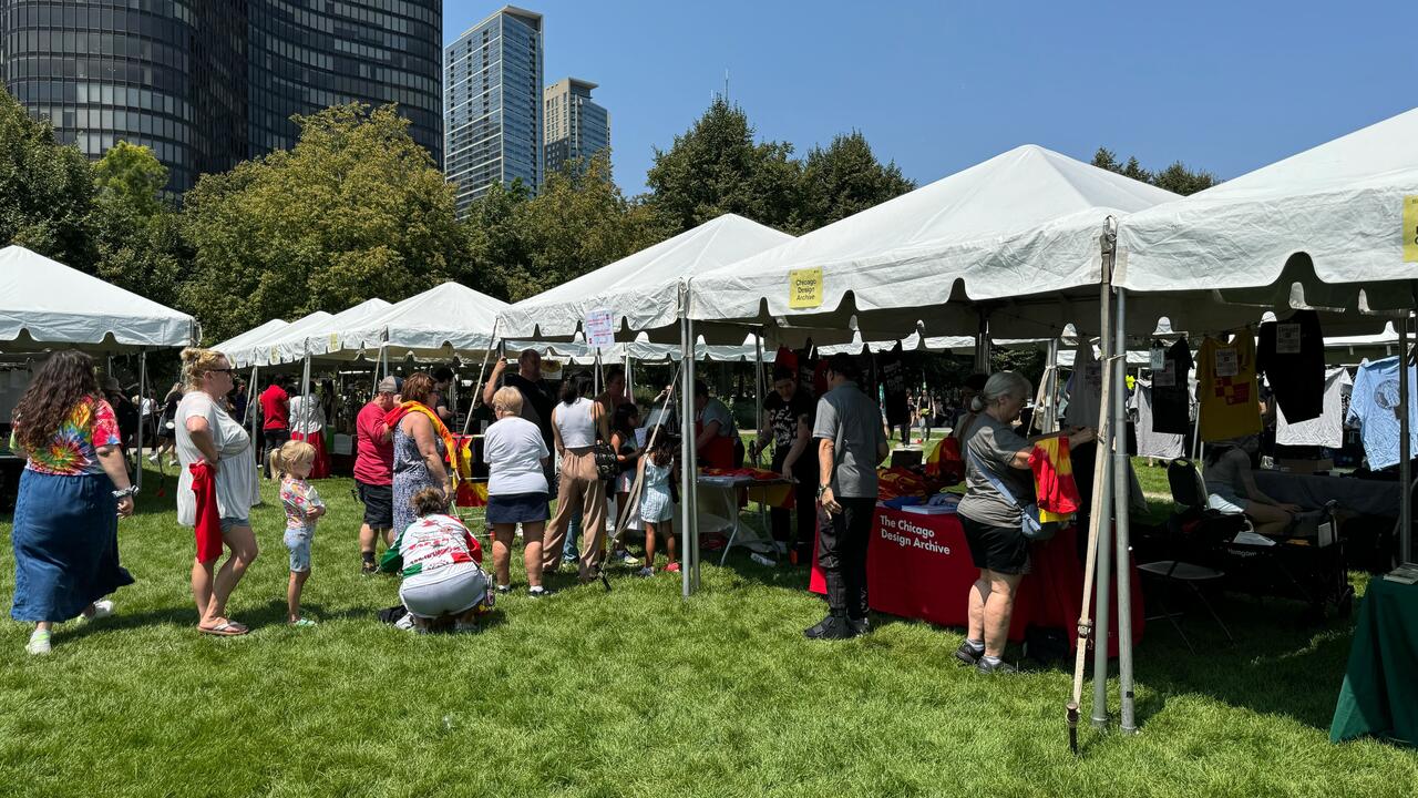 Outdoor event with people browsing stalls under white tents on a grass field. Urban skyscrapers and trees form the background. A sunny day with a clear blue sky.