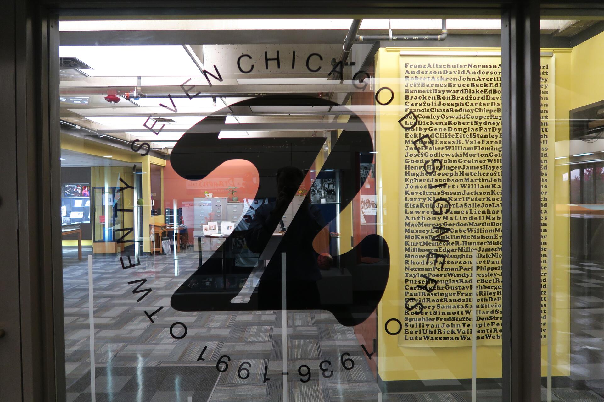 Large "27" logo on glass door, reflecting a hallway with yellow and pink walls. A list of names is visible on the yellow wall in the background.
