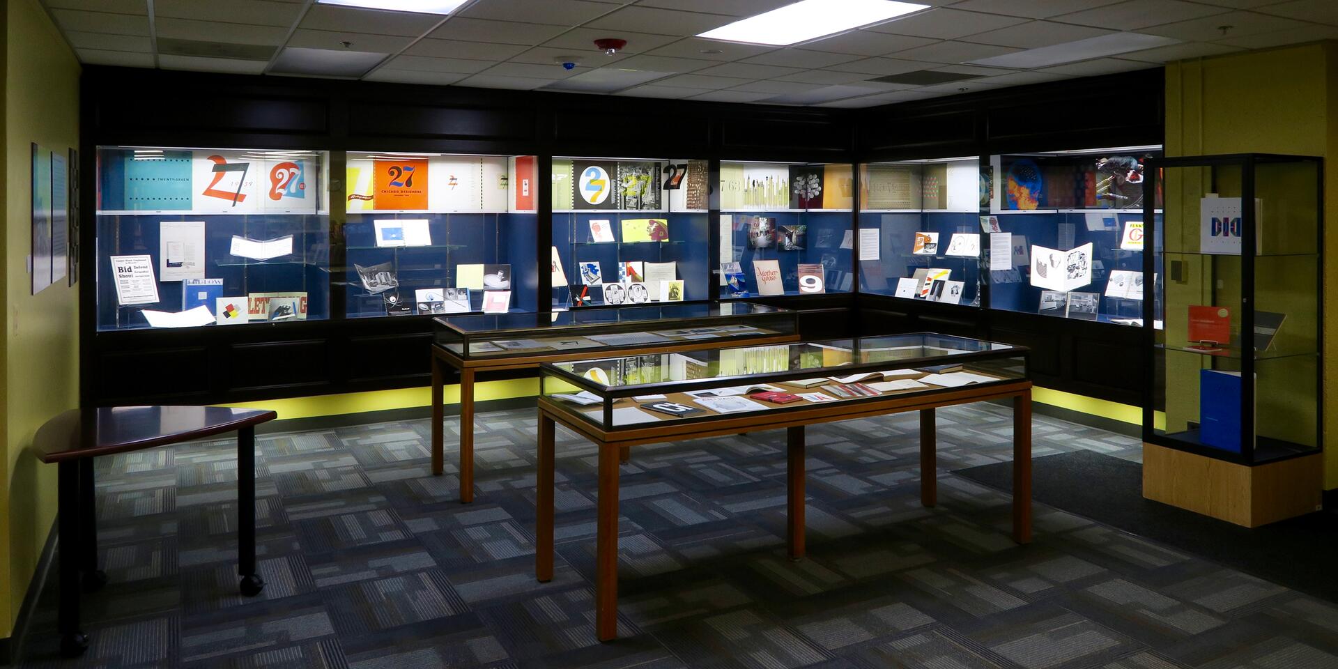 Room with display cases showcasing art and design materials. Two tall glass cases in the background contain colorful posters and art, while two table-height cases display books and documents. The room has a tiled floor and fluorescent lighting.