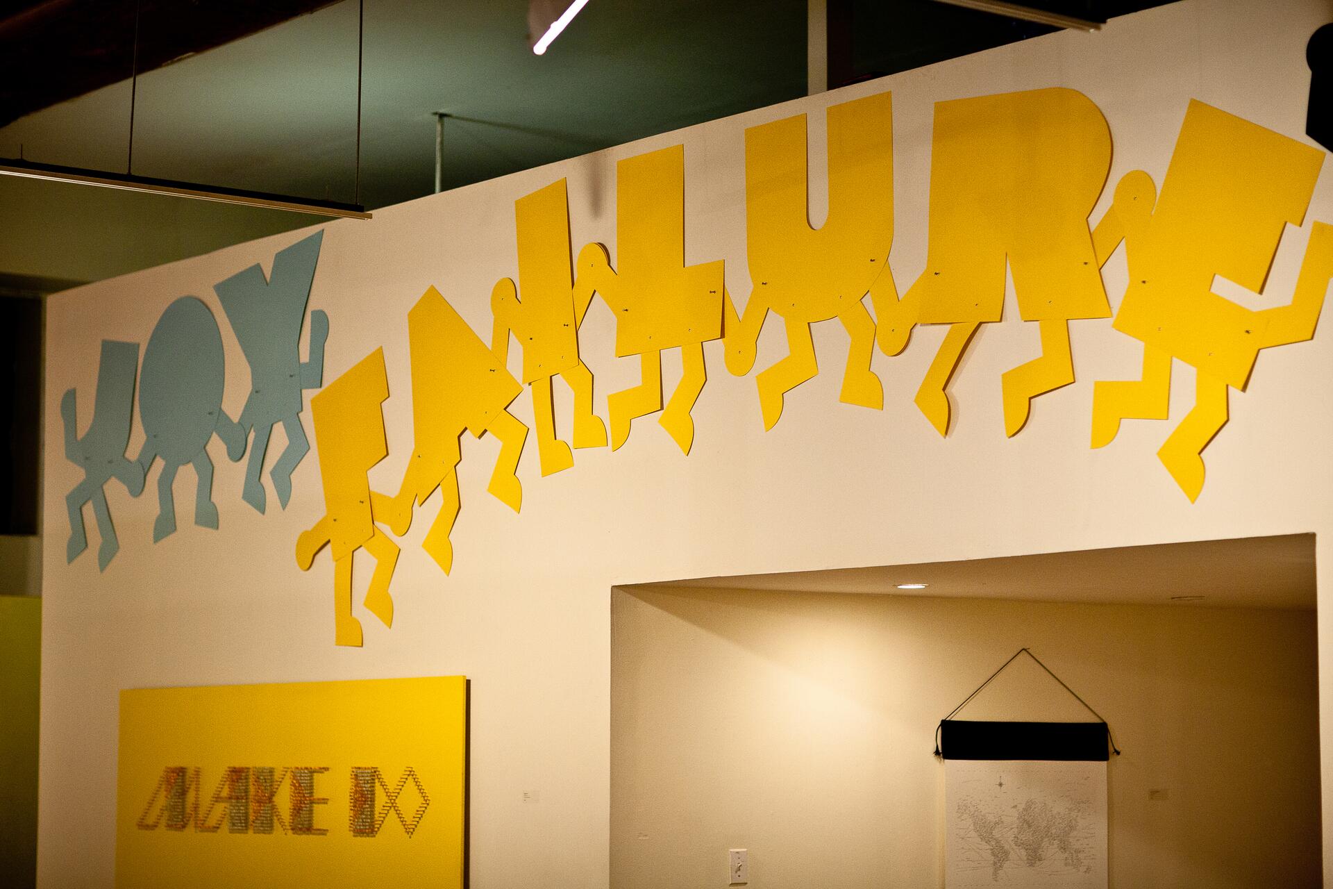 Colorful wall art with stylized figures spelling "JOY FAILURE" in blue and yellow letters. Below, a yellow rectangle displays fragmented text. The setting is an indoor gallery with muted lighting.