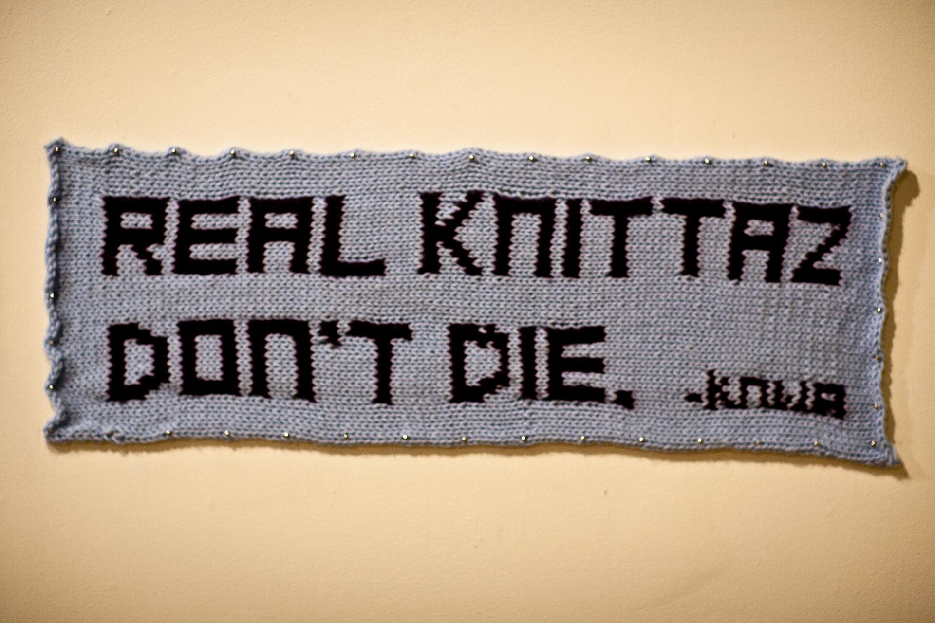 Rectangular knitted wall art with the text "REAL KNITTAZ DON'T DIE" in black lettering on a light gray background.