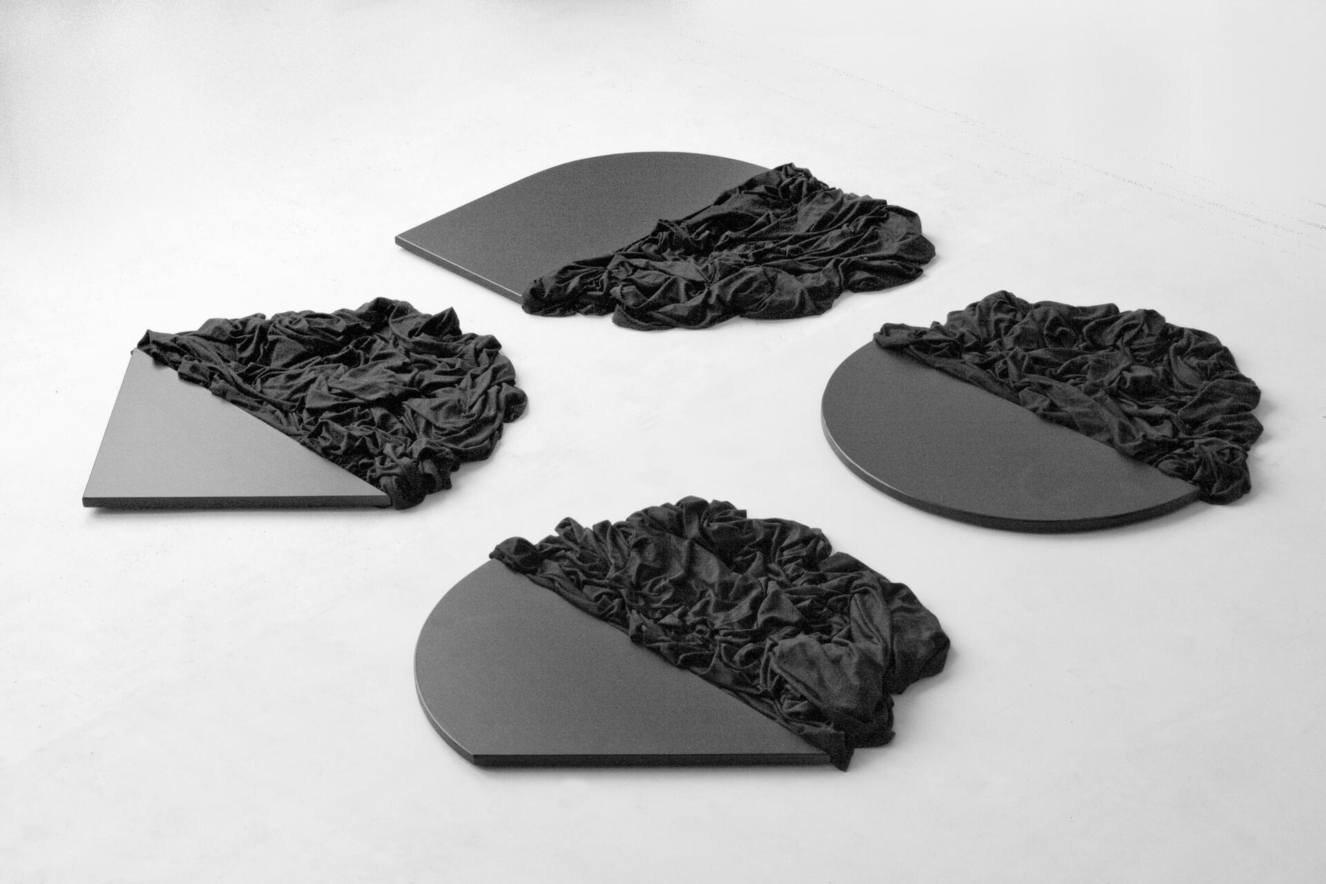 Four flat geometric black shapes, each partially covered with crumpled black fabric, arranged on a white floor.