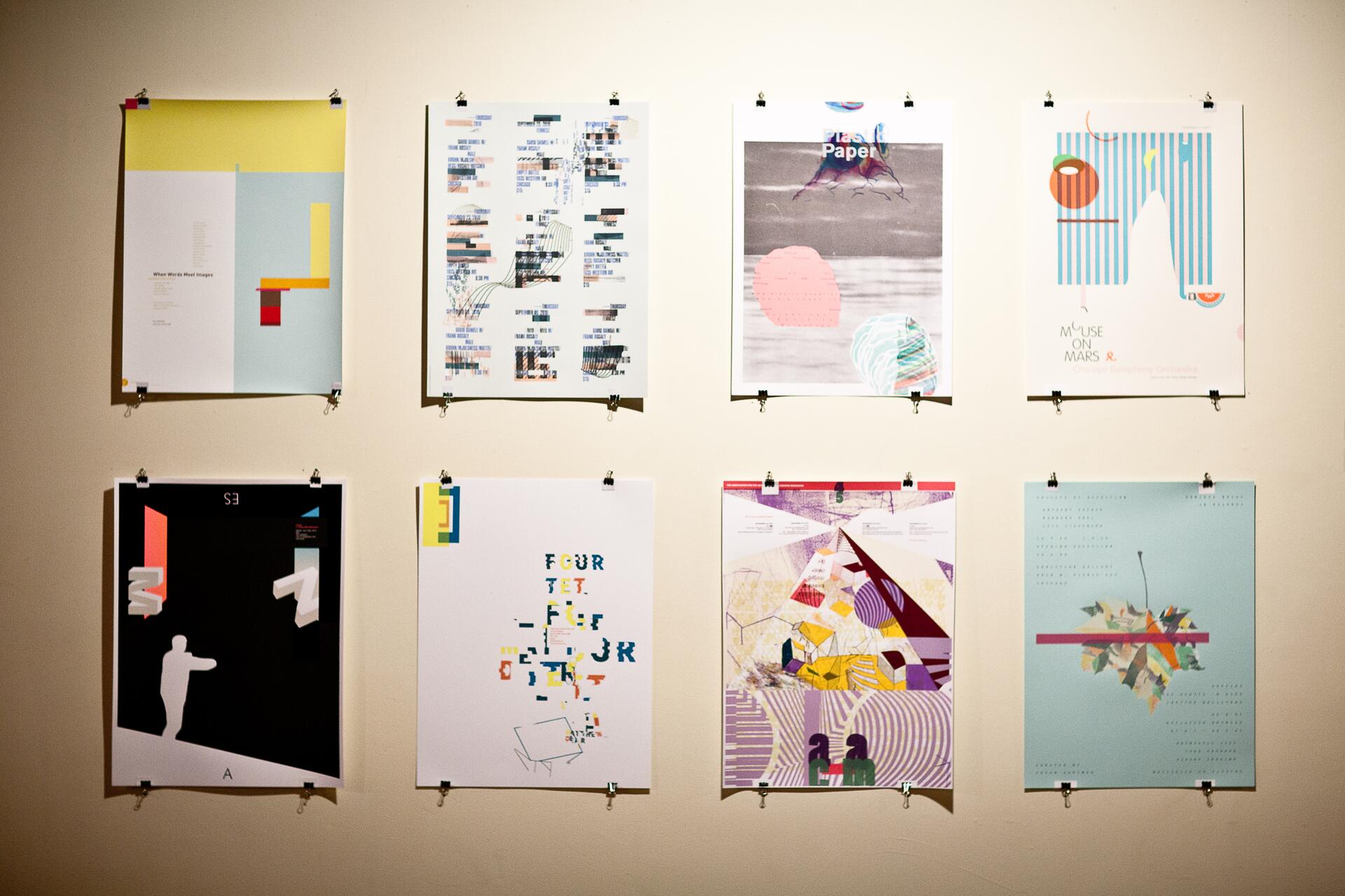 Eight colorful abstract art posters clipped to a beige wall, featuring geometric shapes, text, and mixed media designs, arranged in two rows of four.