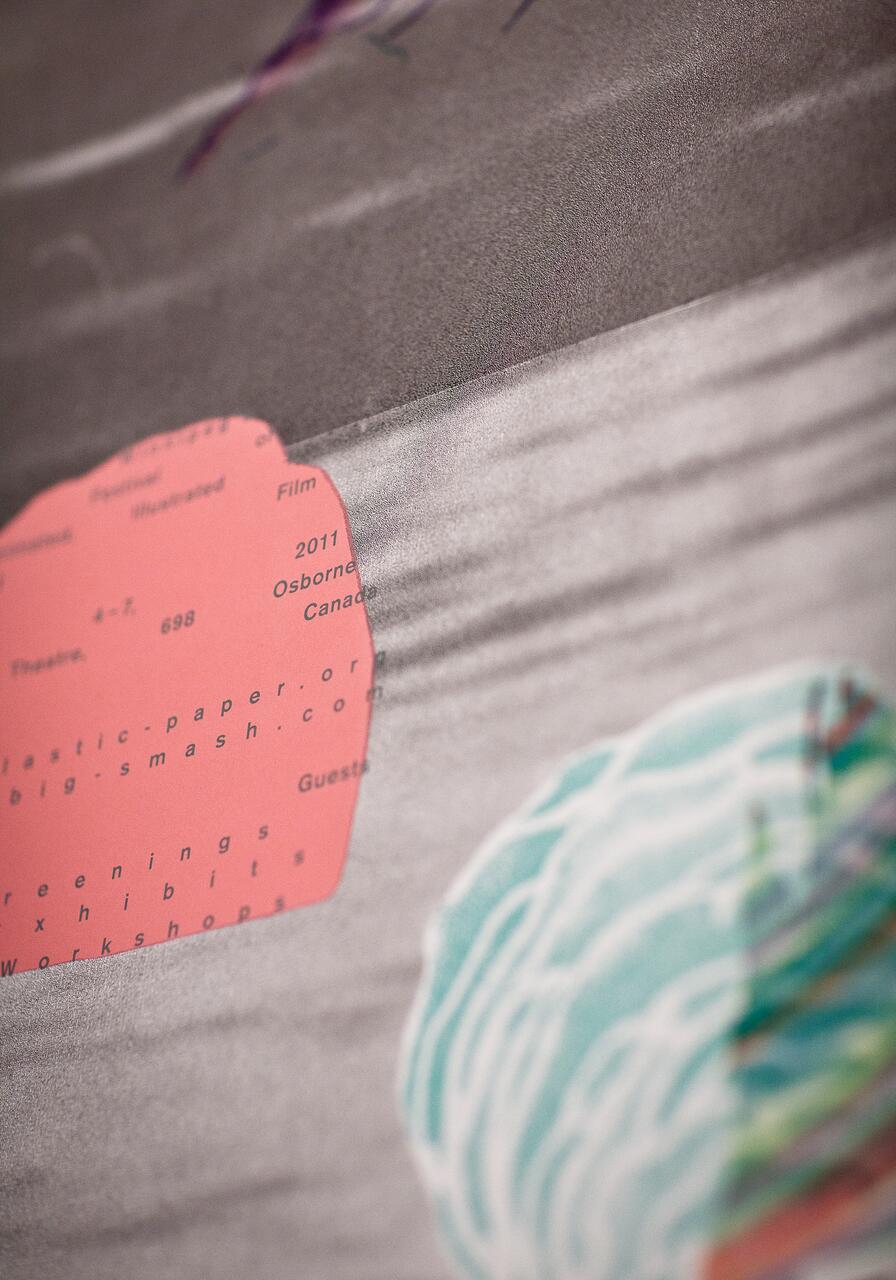 Close-up of a textured surface with a pink oval containing small text, partially visible against a grayish background. A wavy teal design appears at the bottom right.