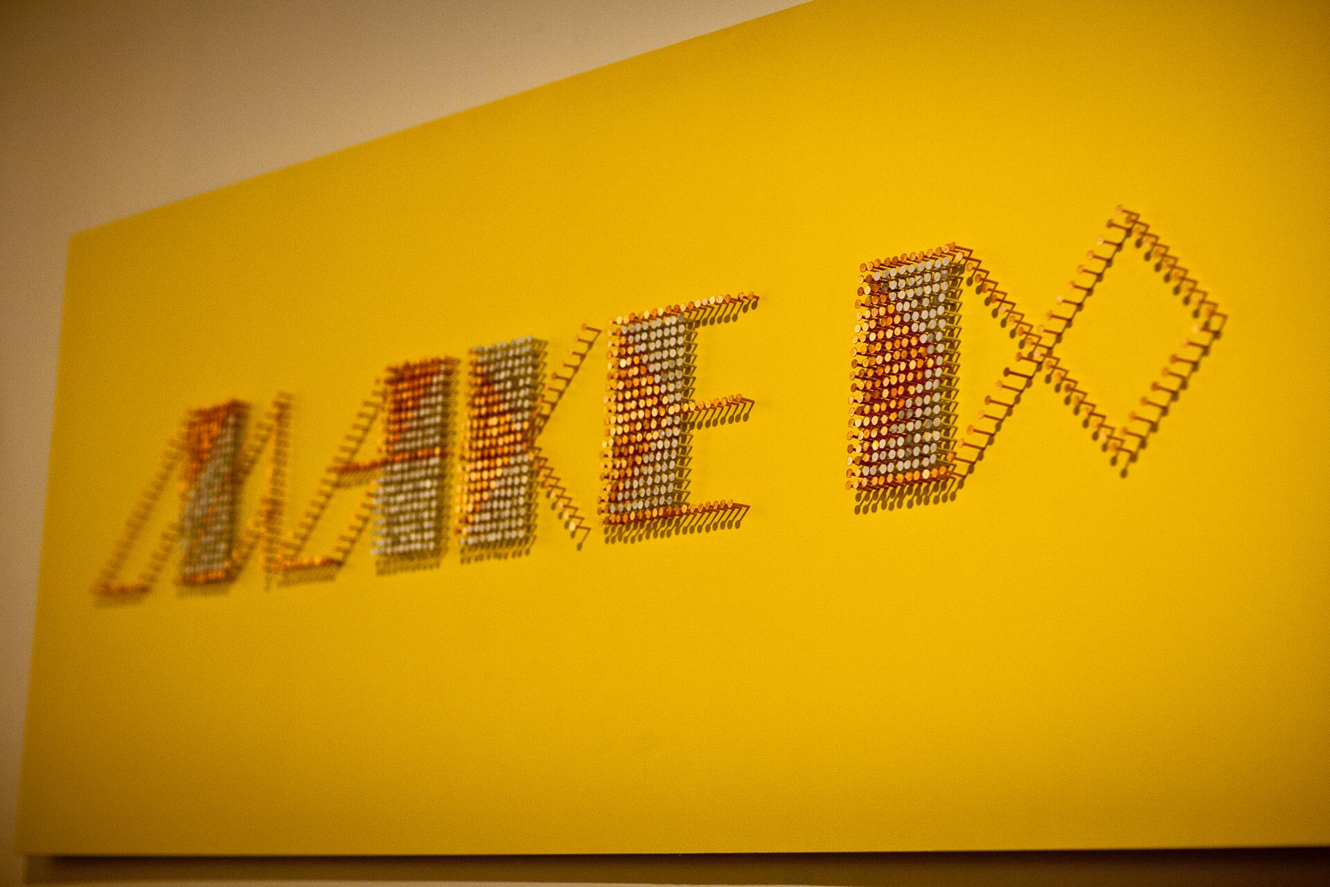 Colorful letters made from numerous small objects on a bright yellow wall, forming the word "MAKE".