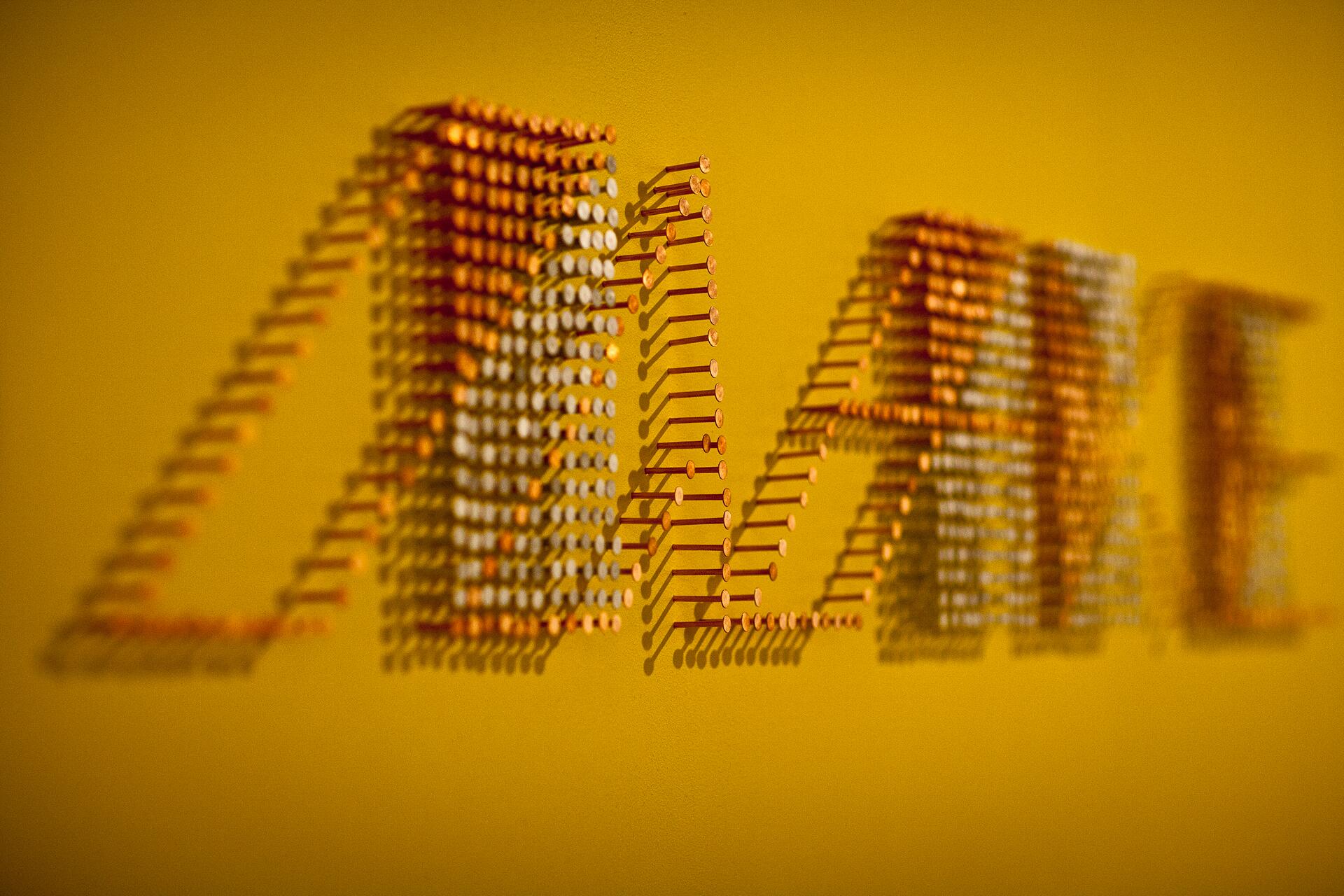 The word "MAKE" is formed using long nails protruding from a yellow wall, casting shadows that enhance the three-dimensional effect.