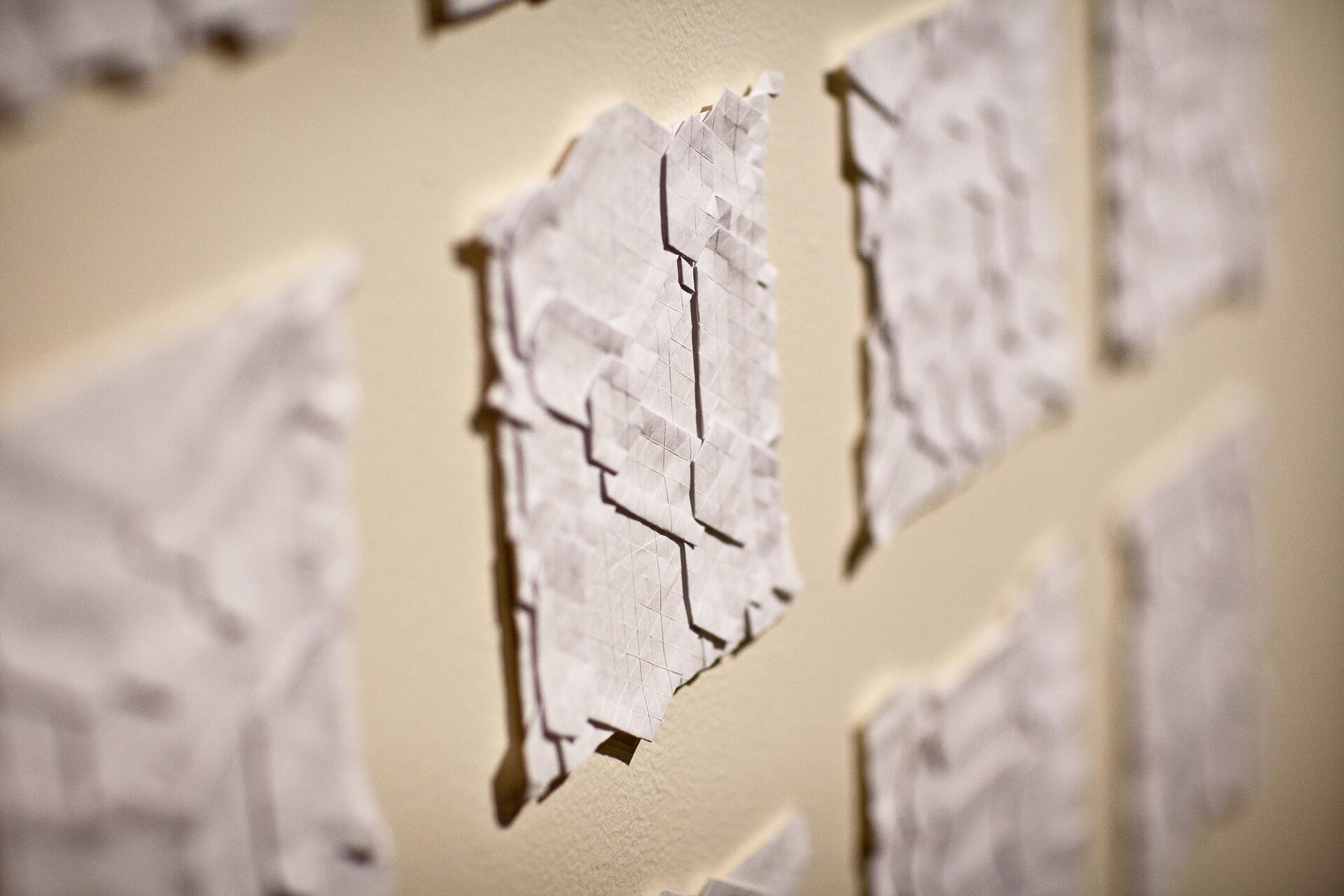 Geometric folded paper art displayed on a wall, with soft lighting and shadows enhancing the texture.