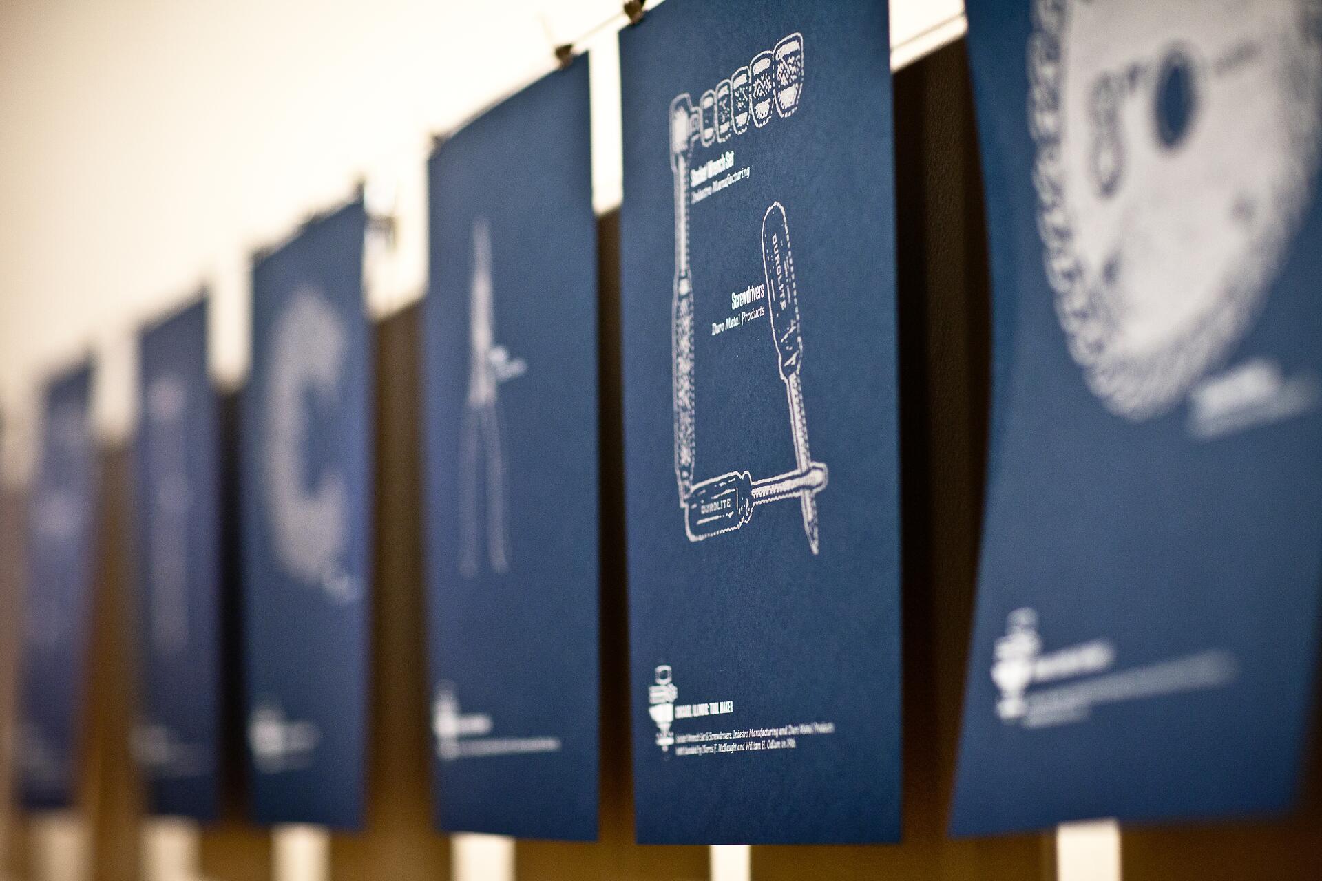 Blue posters with white technical drawings hanging in a row, showing tools like a screwdriver. The focus is on the central poster, while others are blurred in the background.