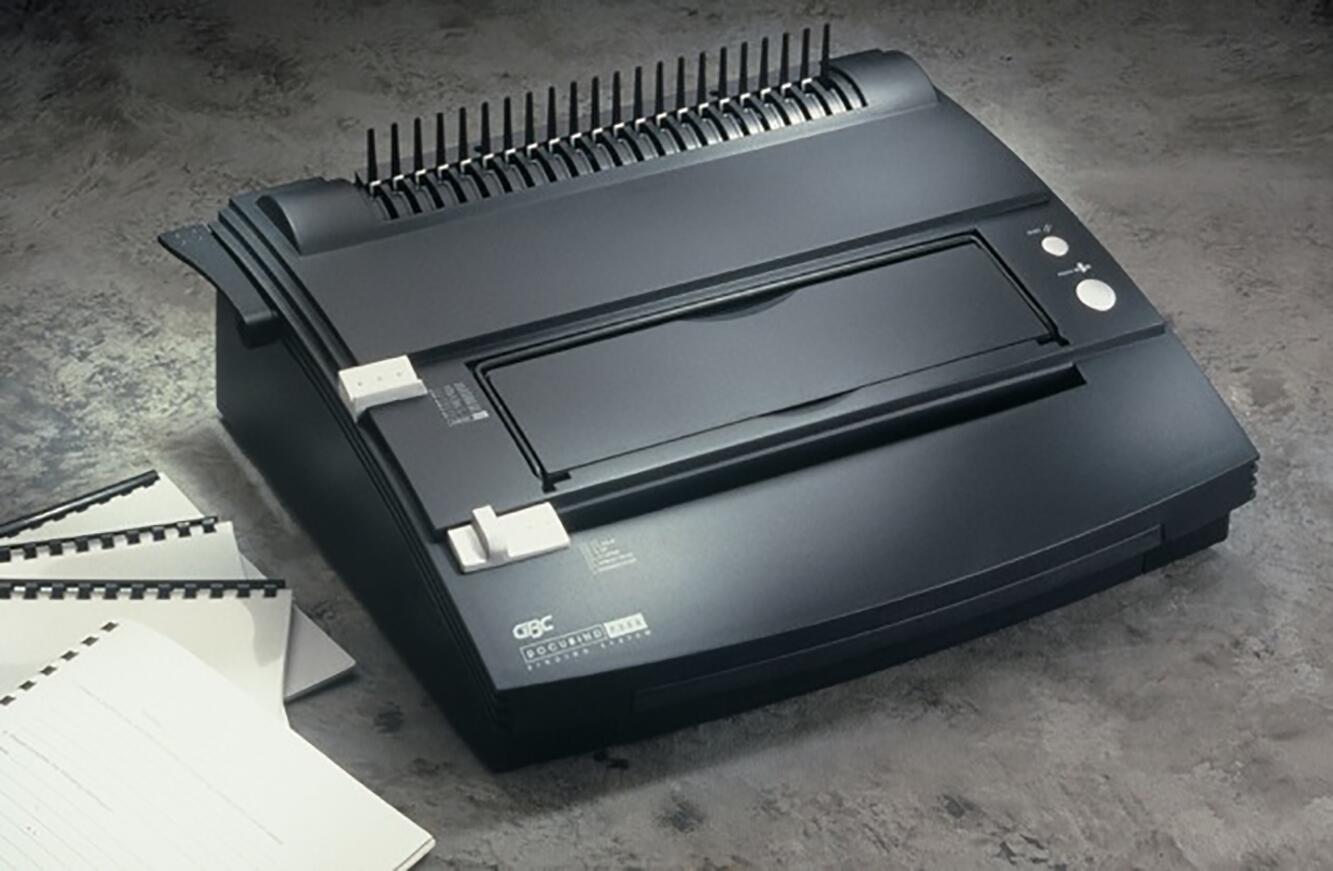 Black comb binding machine on a textured surface, with three spiral-bound booklets nearby.