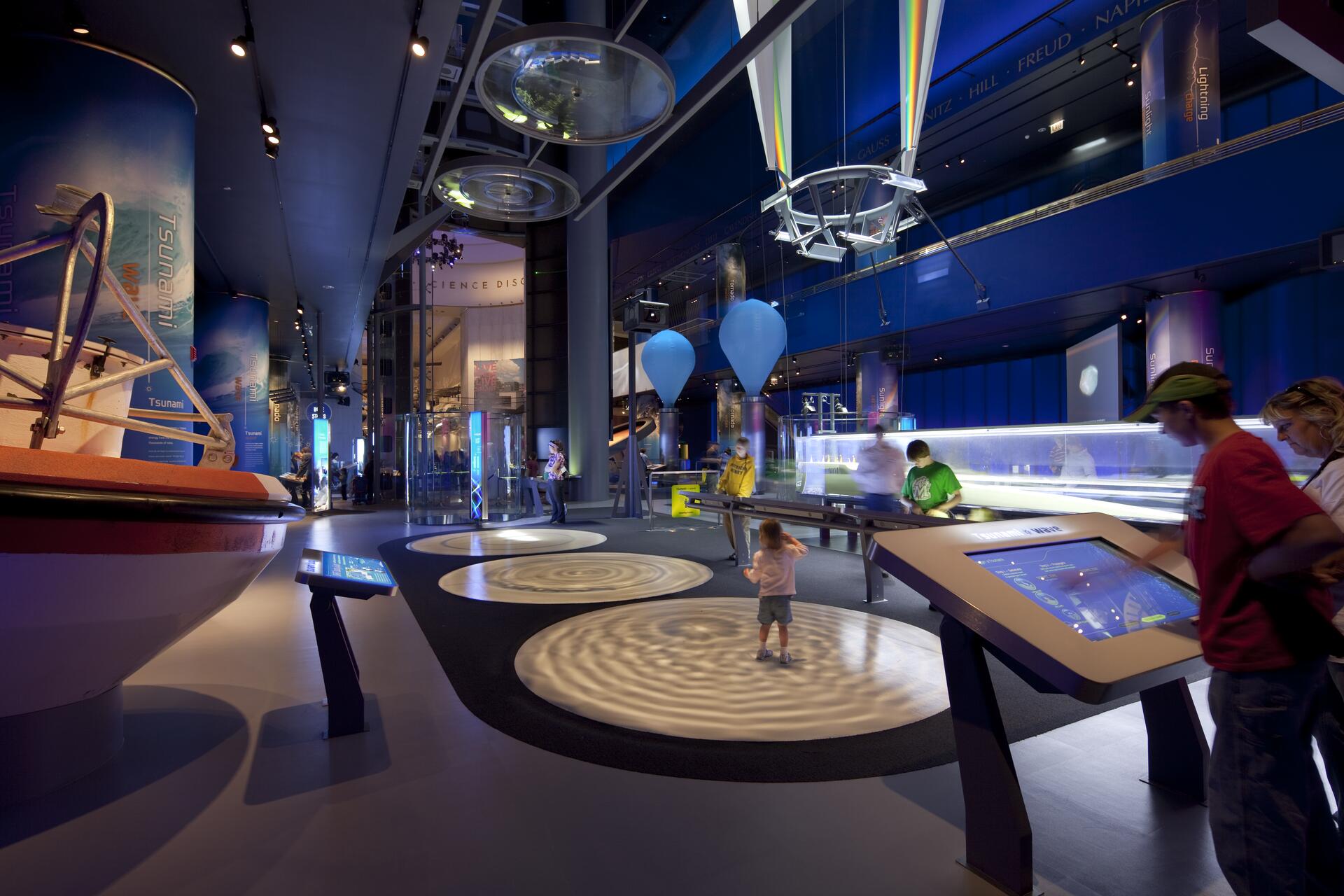 Interactive science museum exhibit with large cylindrical displays, projections on the floor, and visitors engaging with digital screens. The space is dimly lit with blue and green lighting, creating a futuristic ambiance.