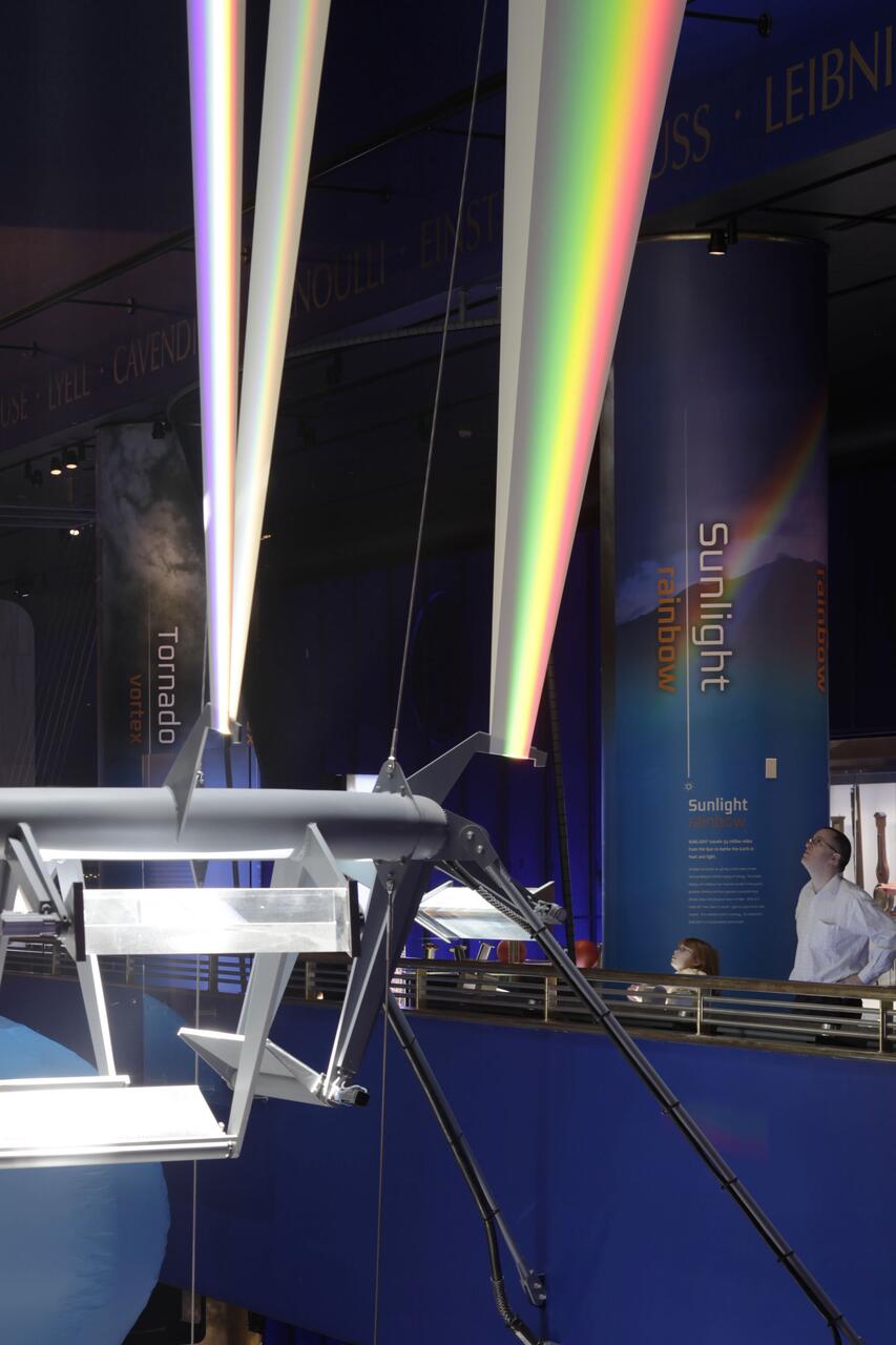 Exhibit featuring a large apparatus reflecting colorful rainbow light beams in a dark room, with people observing from a railing.