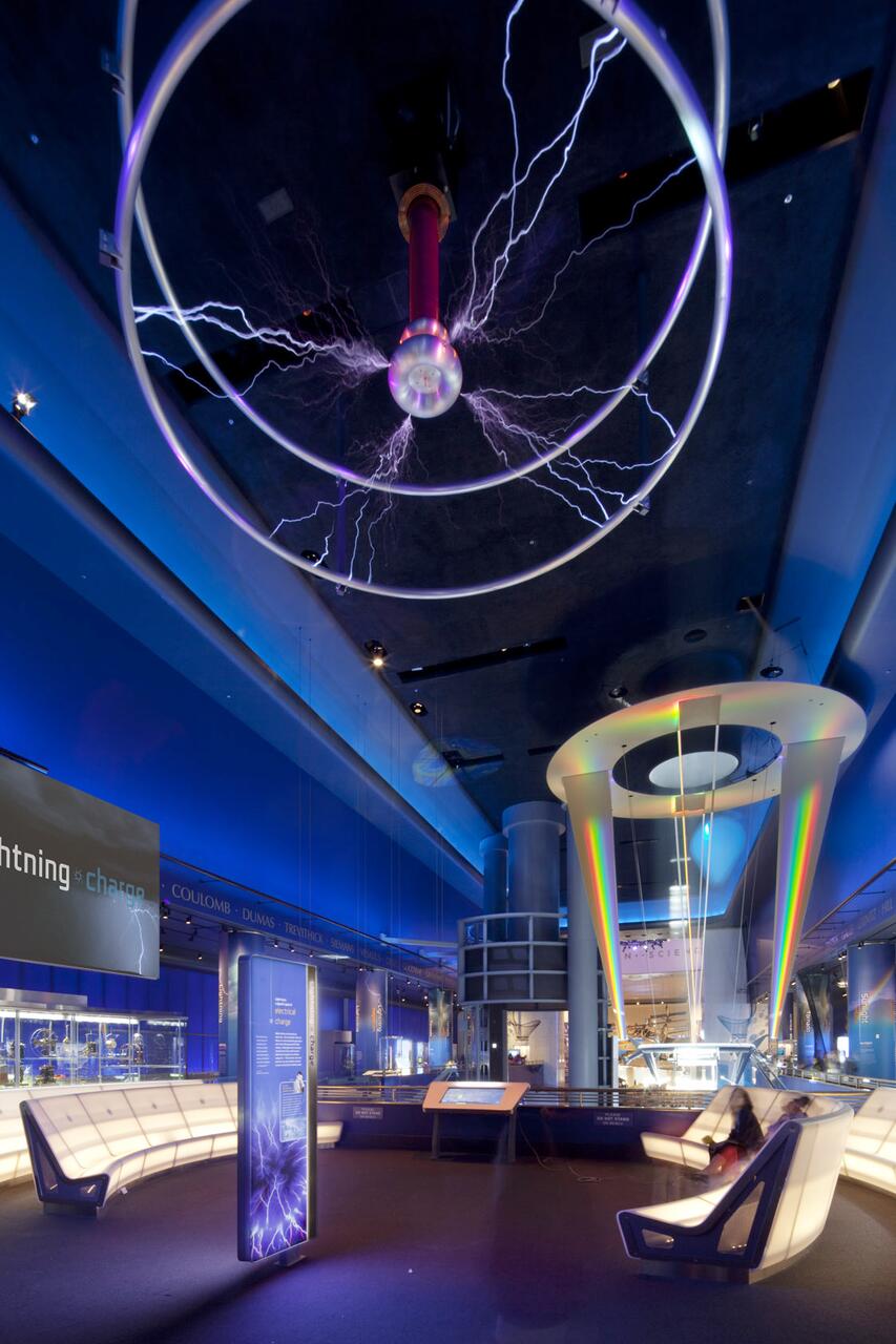 Dark room in a science museum with a Tesla coil sparking electricity, surrounded by exhibits and illuminated blue lighting.