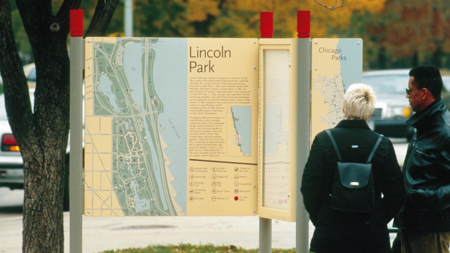 Two people stand in front of outdoor informational boards about Lincoln Park and Chicago Parks, featuring maps and text. Autumn foliage and a parked car are visible in the background.