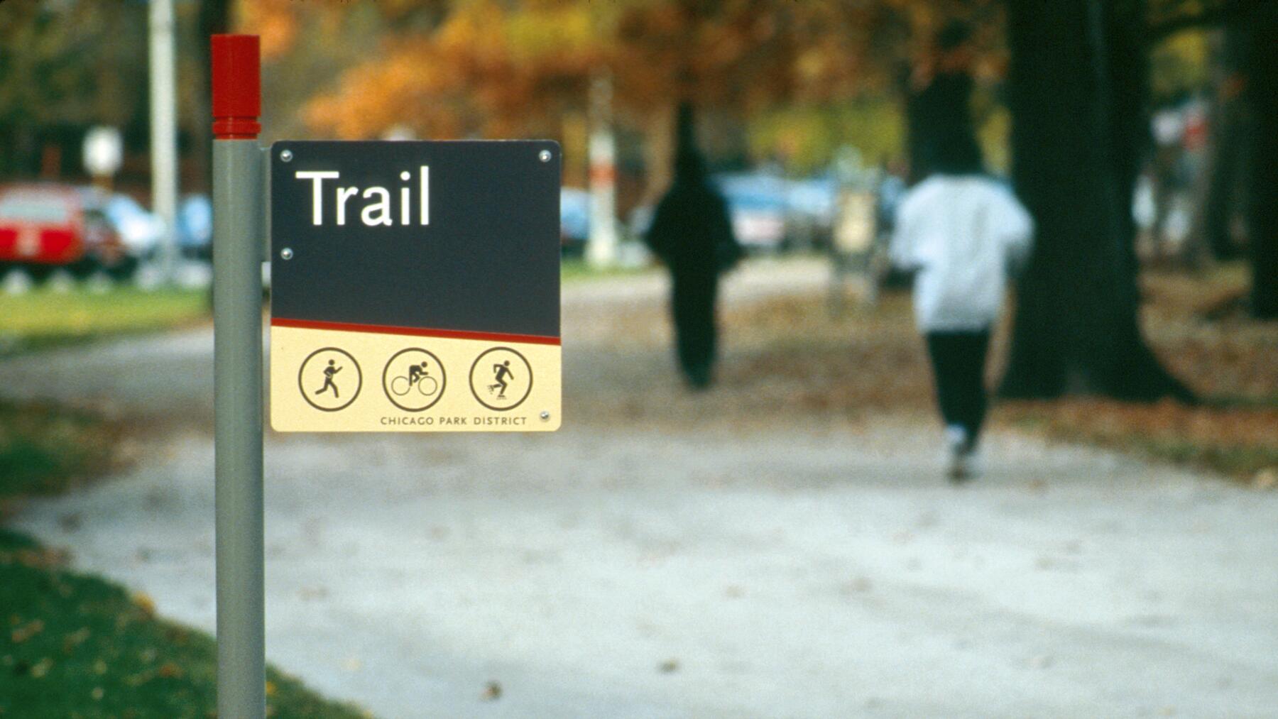 Trail sign in a park with autumn leaves and two people walking on a path.