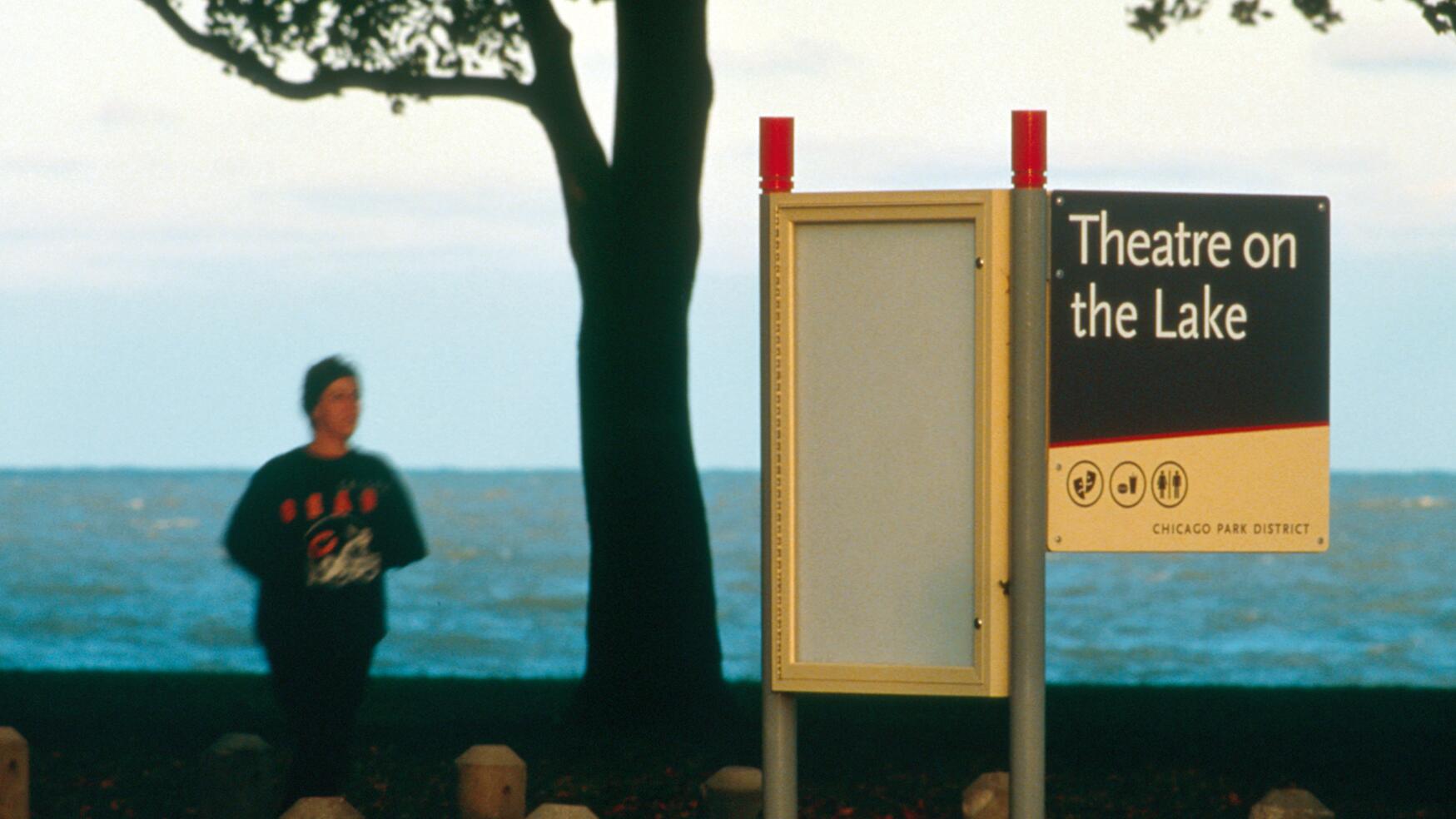 Sign for "Theatre on the Lake" by a waterfront with a person jogging in the background.