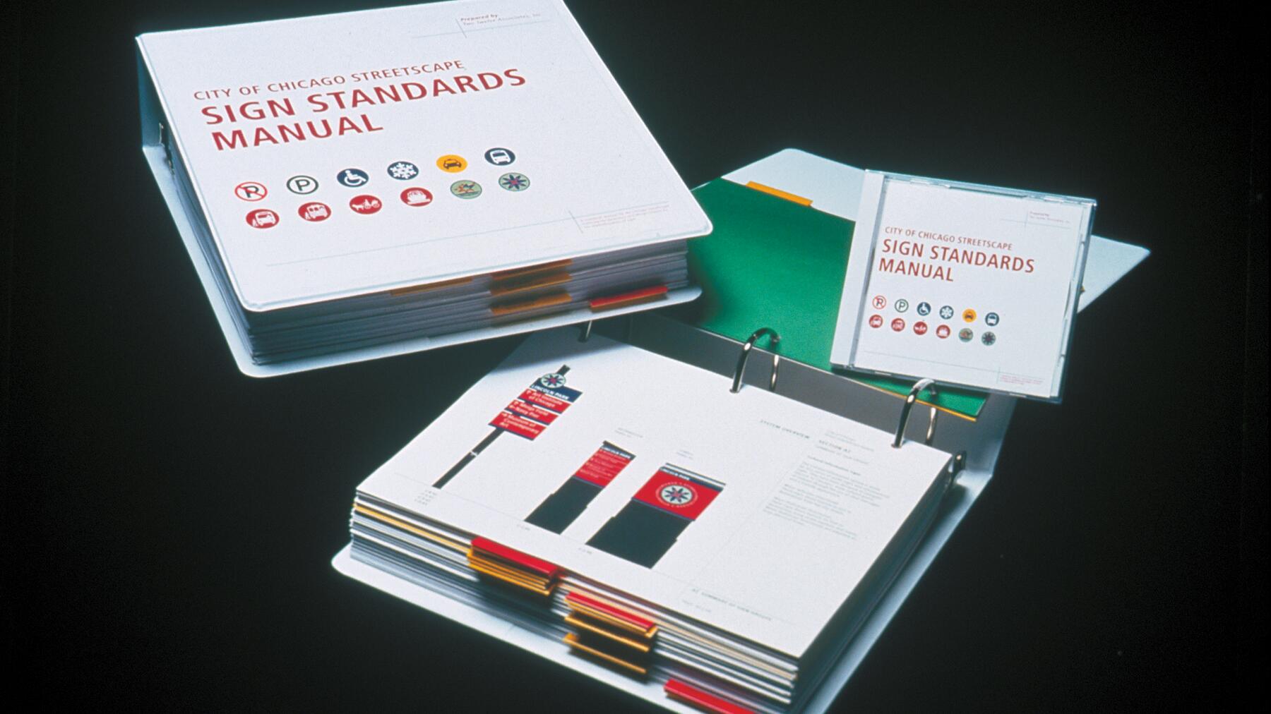 Open binder with pages showing street sign standards, colorful tabs visible. Nearby are two binders stacked and a matching CD case, all labeled "City of Chicago Streetscape Sign Standards Manual," set against a black background.