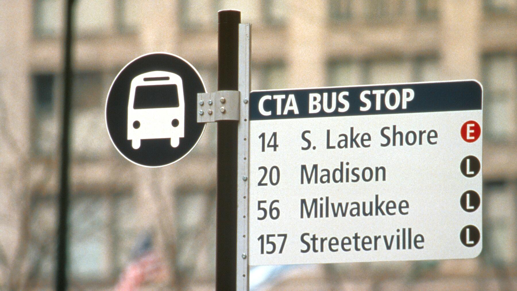 CTA bus stop sign with routes 14, 20, 56, and 157 listed for S. Lake Shore, Madison, Milwaukee, and Streeterville. The background is blurred urban buildings.
