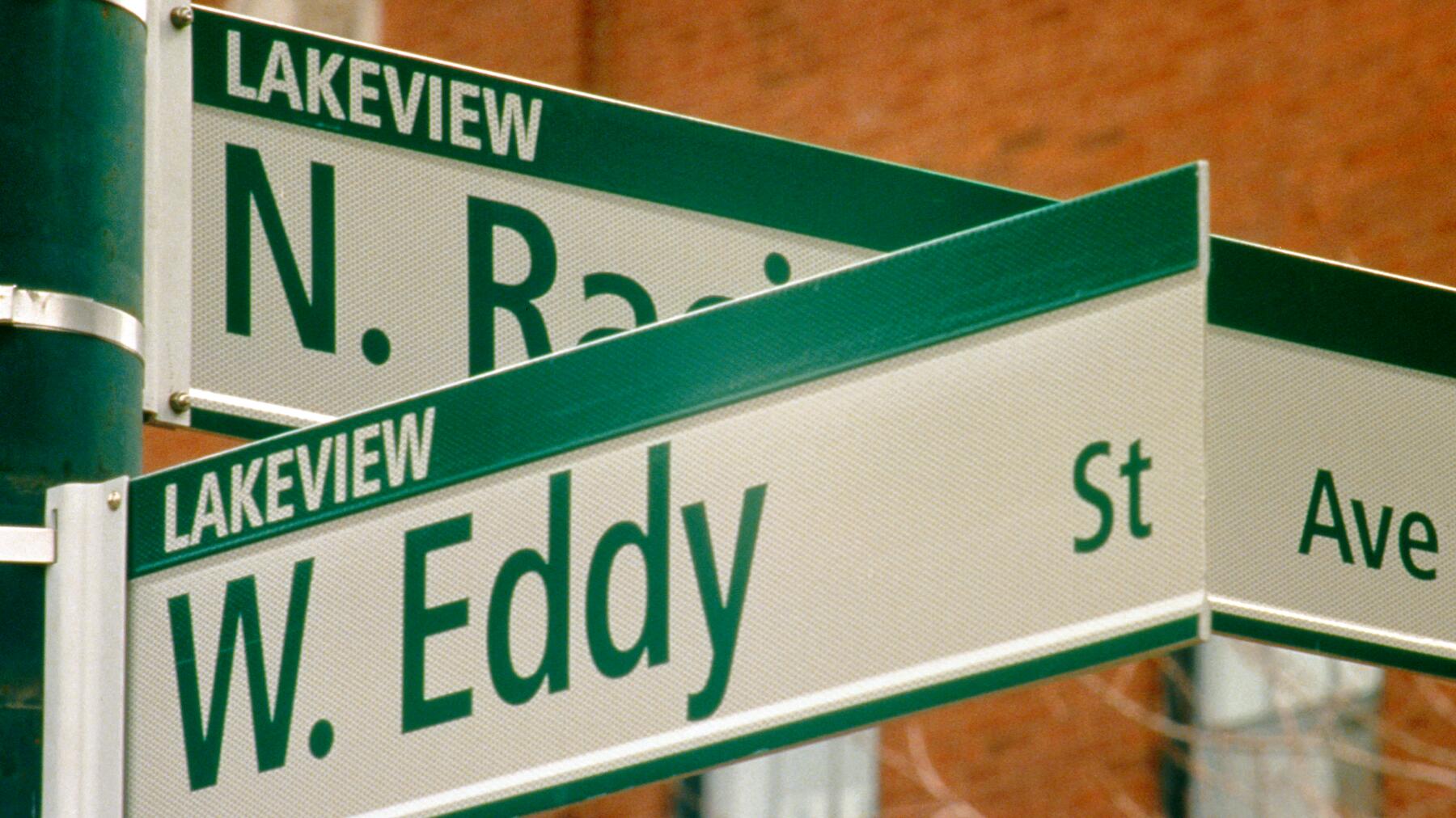 Street signs at an intersection display "N. Raci..." and "W. Eddy" with "Lakeview" labeled above each. A green pole supports the signs against a blurred brick background.