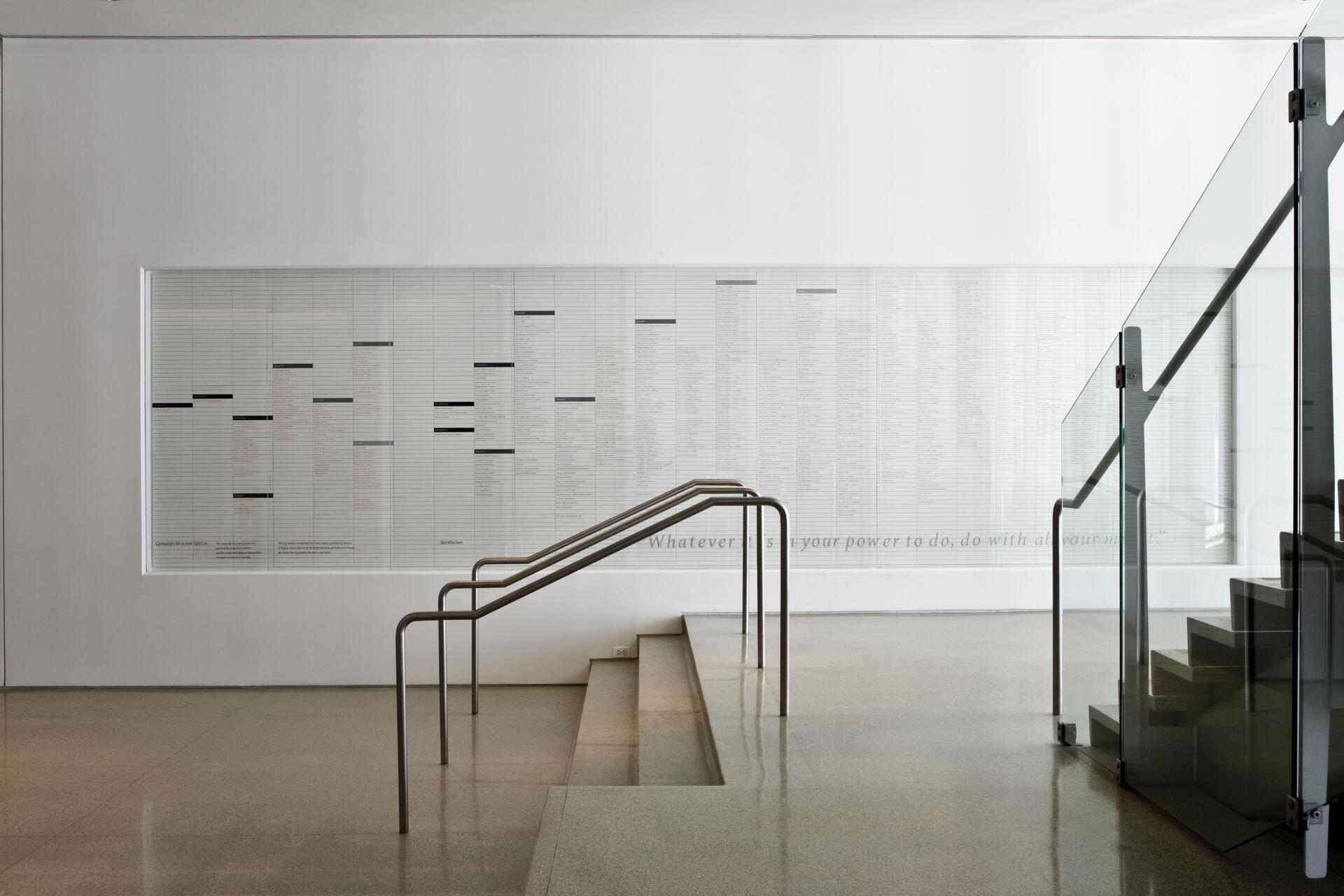 Staircase with sleek metal railings and glass panels in a modern, minimalistic setting. A text-filled white wall in the background, with smooth marble flooring.