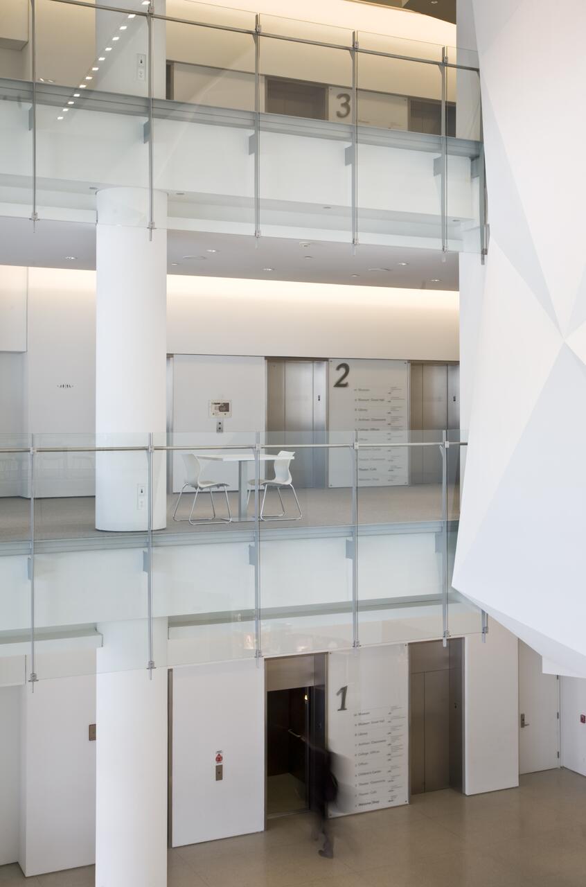 Modern multi-level interior with glass railings, white walls, and elevators marked with numbers 1, 2, and 3. A small table and chairs are visible on the second floor.