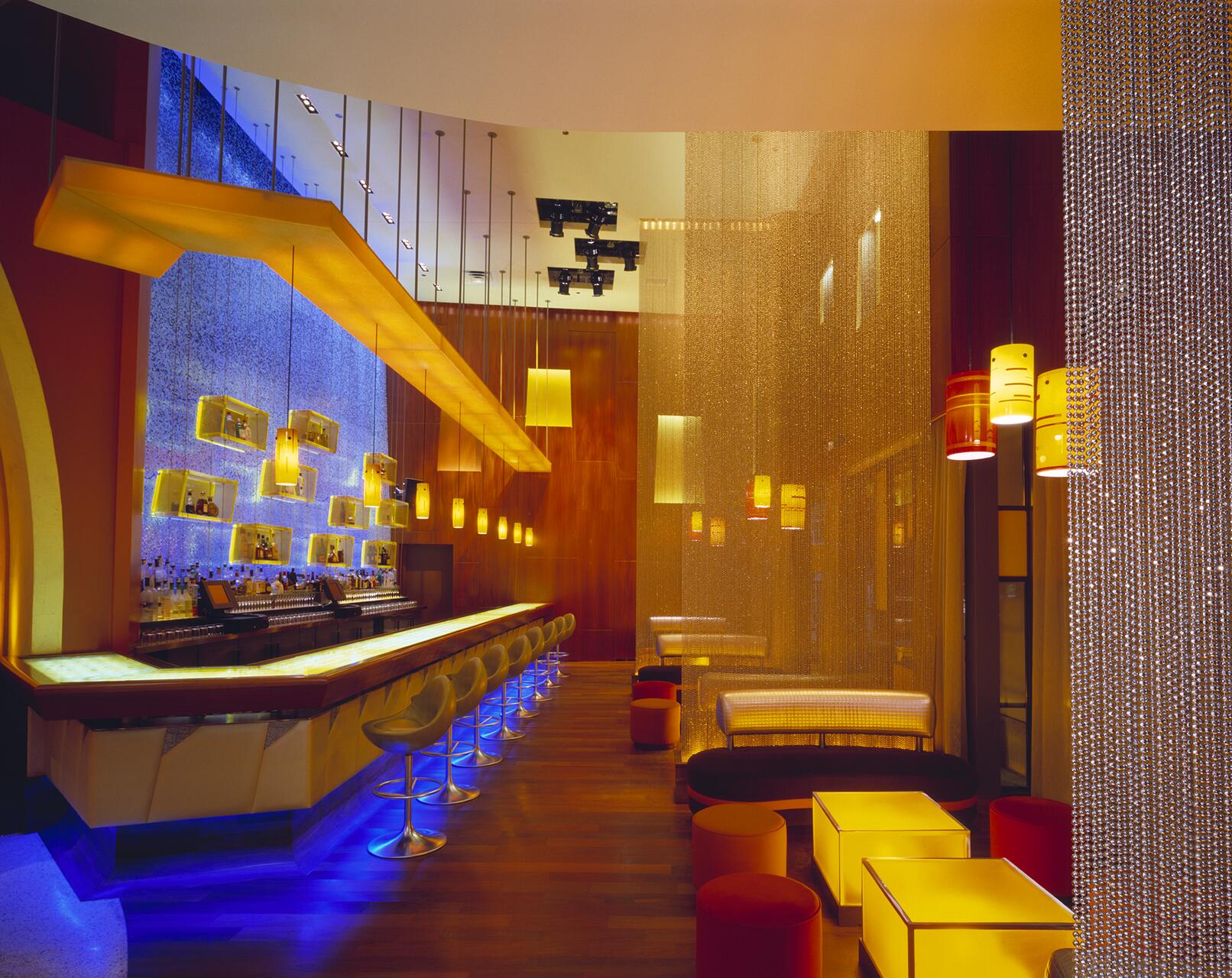 Modern bar interior with vibrant blue and yellow lighting. The bar counter is lined with round stools and illuminated shelves, while small tables and plush seating fill the lounge area. A beaded curtain separates the spaces, enhancing the elegant ambiance.