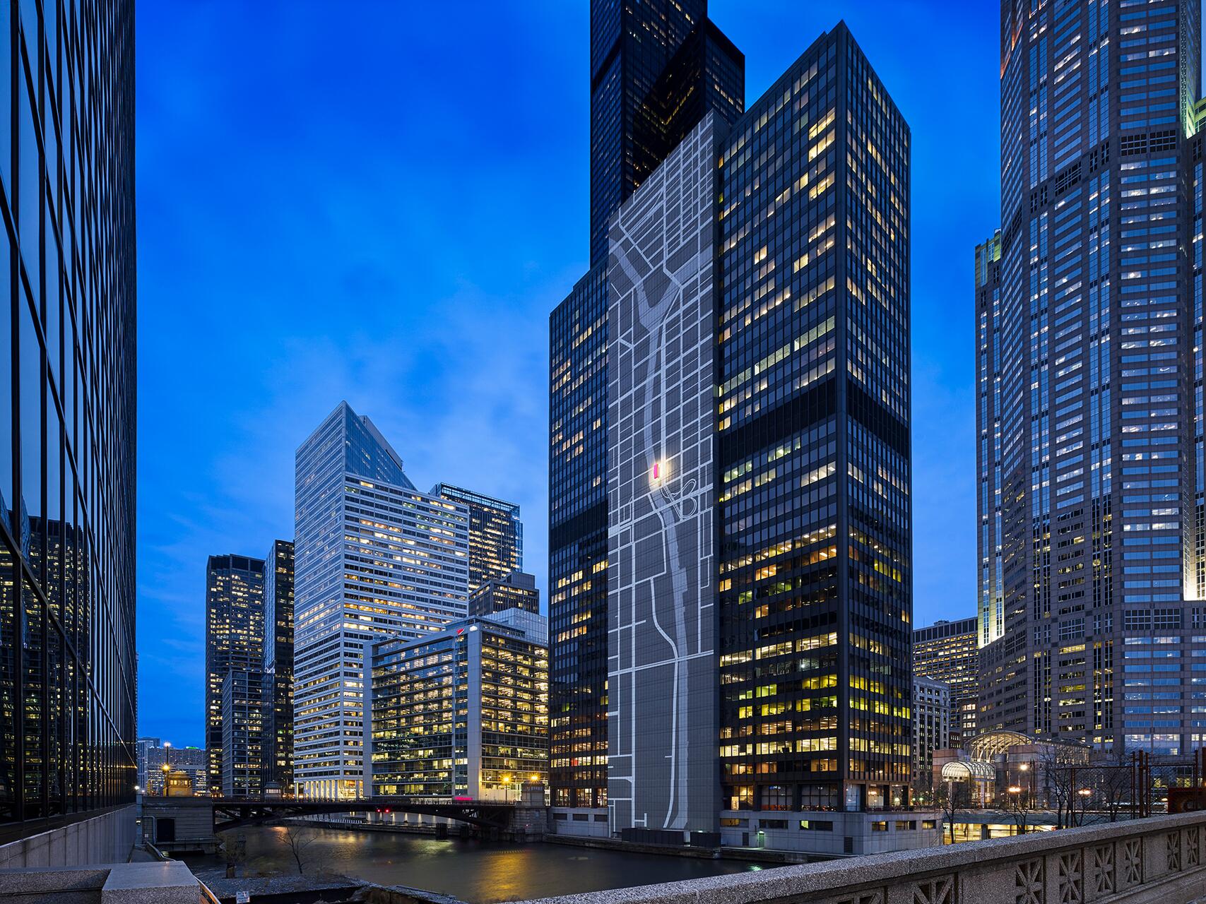 Cityscape at dusk with tall illuminated skyscrapers reflecting in a river. One building has a large map-like design projected on its facade. The sky is a deep blue, adding contrast to the lit windows.