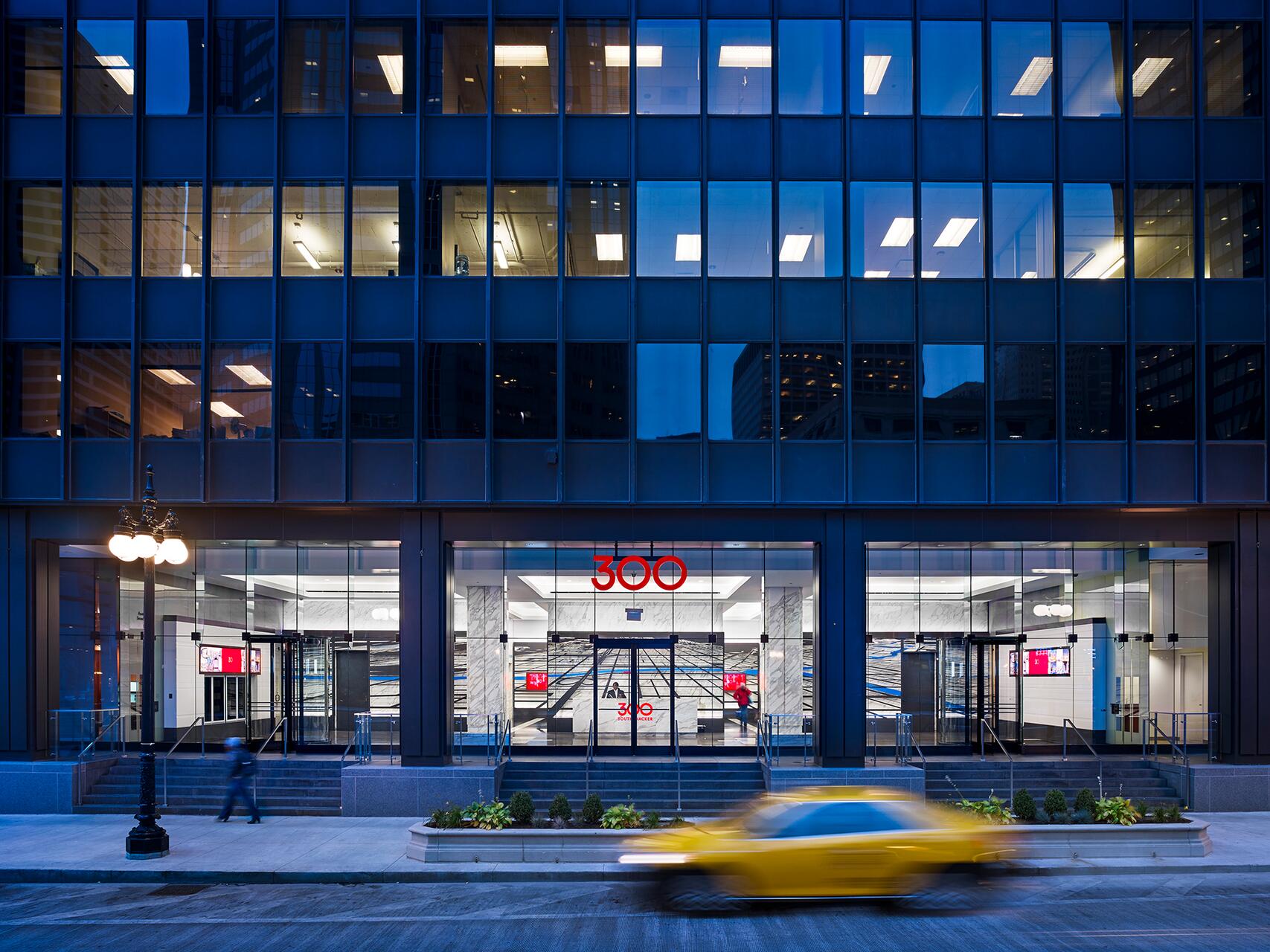 A modern office building with a blue-tinted glass facade at dusk. A walking person and a blurred yellow taxi are in front, and a red "300" is visible above the entrance. Illuminated interior and streetlights enhance the scene.