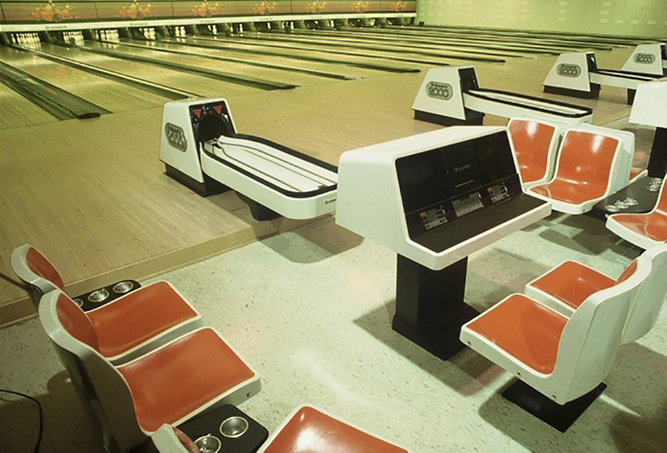 Bowling alley with polished wooden lanes, red and white plastic seating, and electronic scoring consoles.