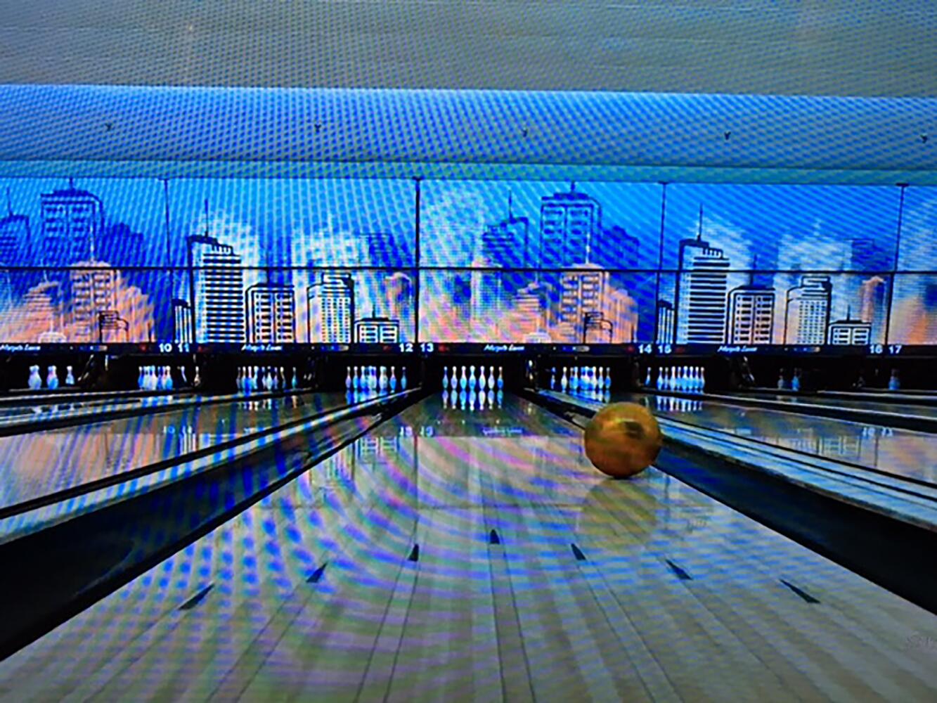 A bowling alley with colorful skyline-themed panels in the background. A golden bowling ball rolls down a lane toward a set of white pins.