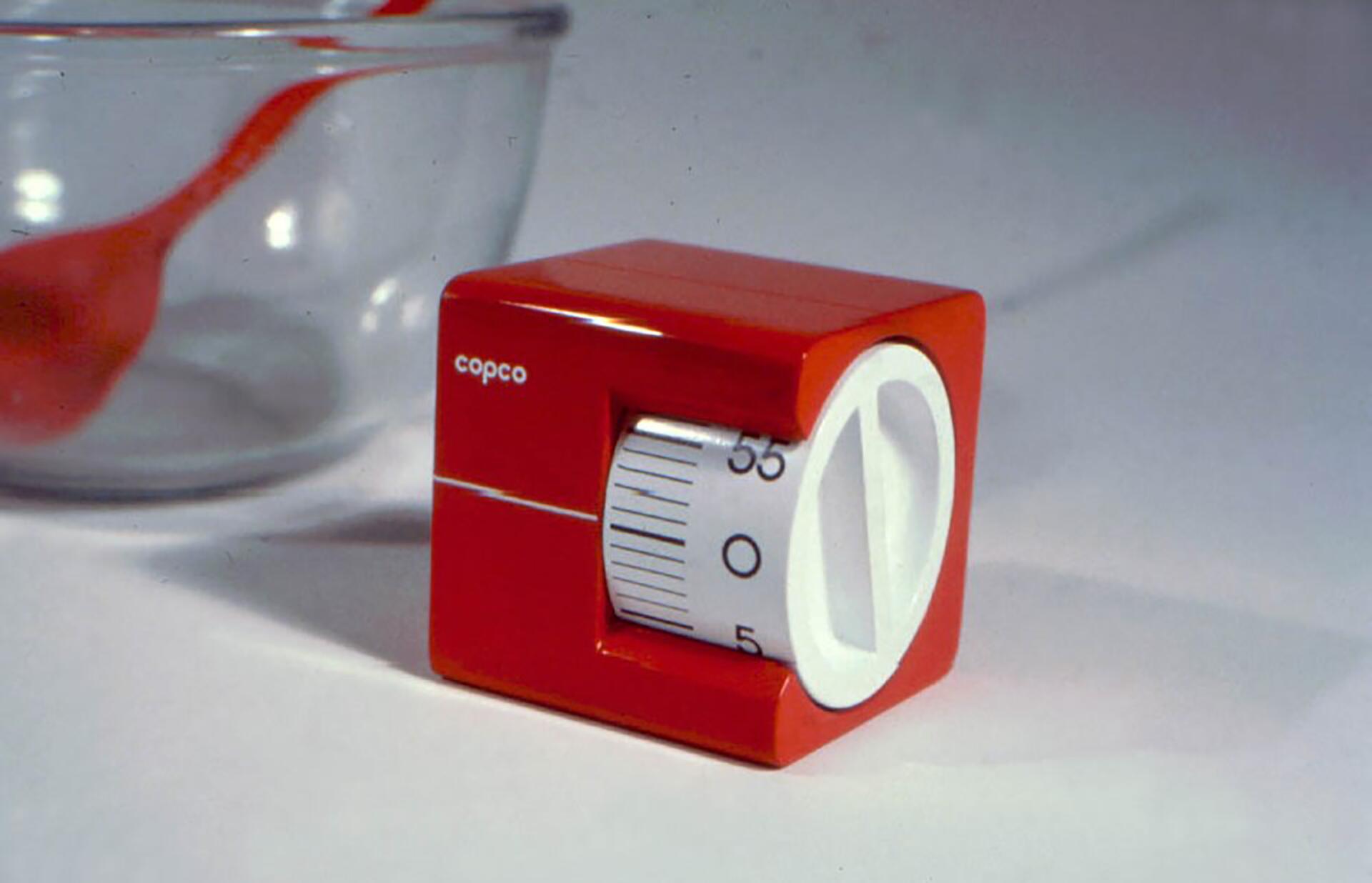 Red kitchen timer with a white dial, set near a clear glass bowl with a red spoon inside, on a light surface.