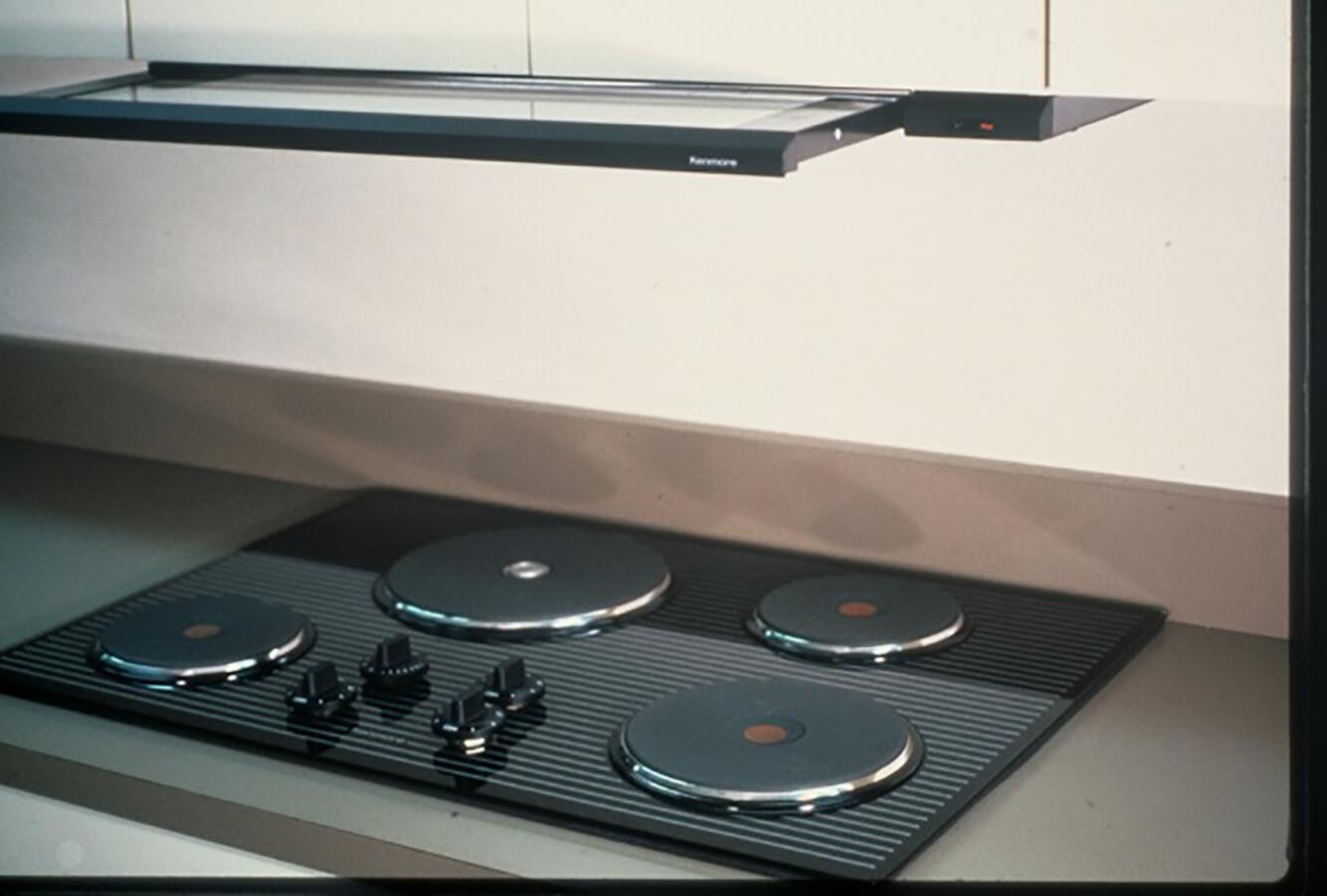 Stovetop with four burners and control knobs, set into a countertop beneath a range hood. The surface is sleek with a modern design, featuring smooth, rounded elements.
