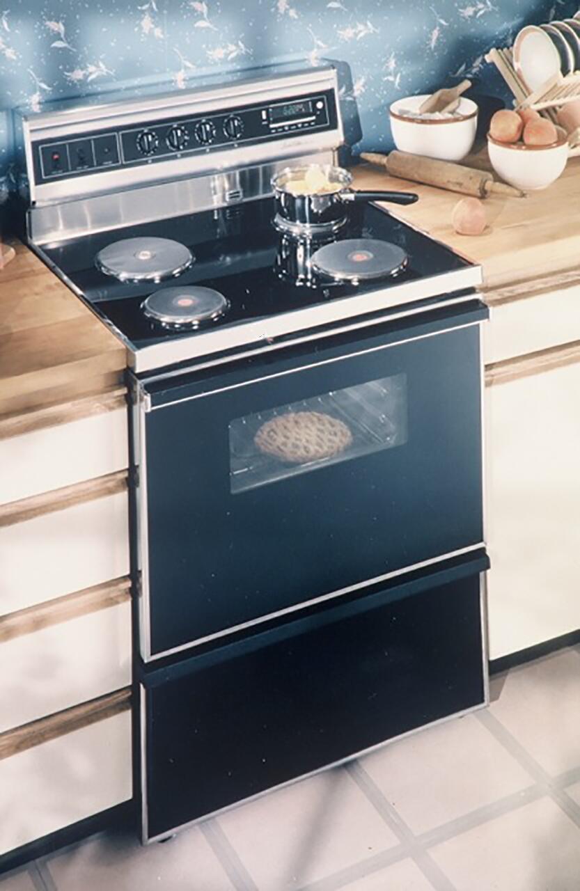 Electric stove with four burners and an oven, featuring a pie inside. A pot with ingredients sits on top. The kitchen counter holds baking items and a bowl of fruit, with floral blue wallpaper in the background.