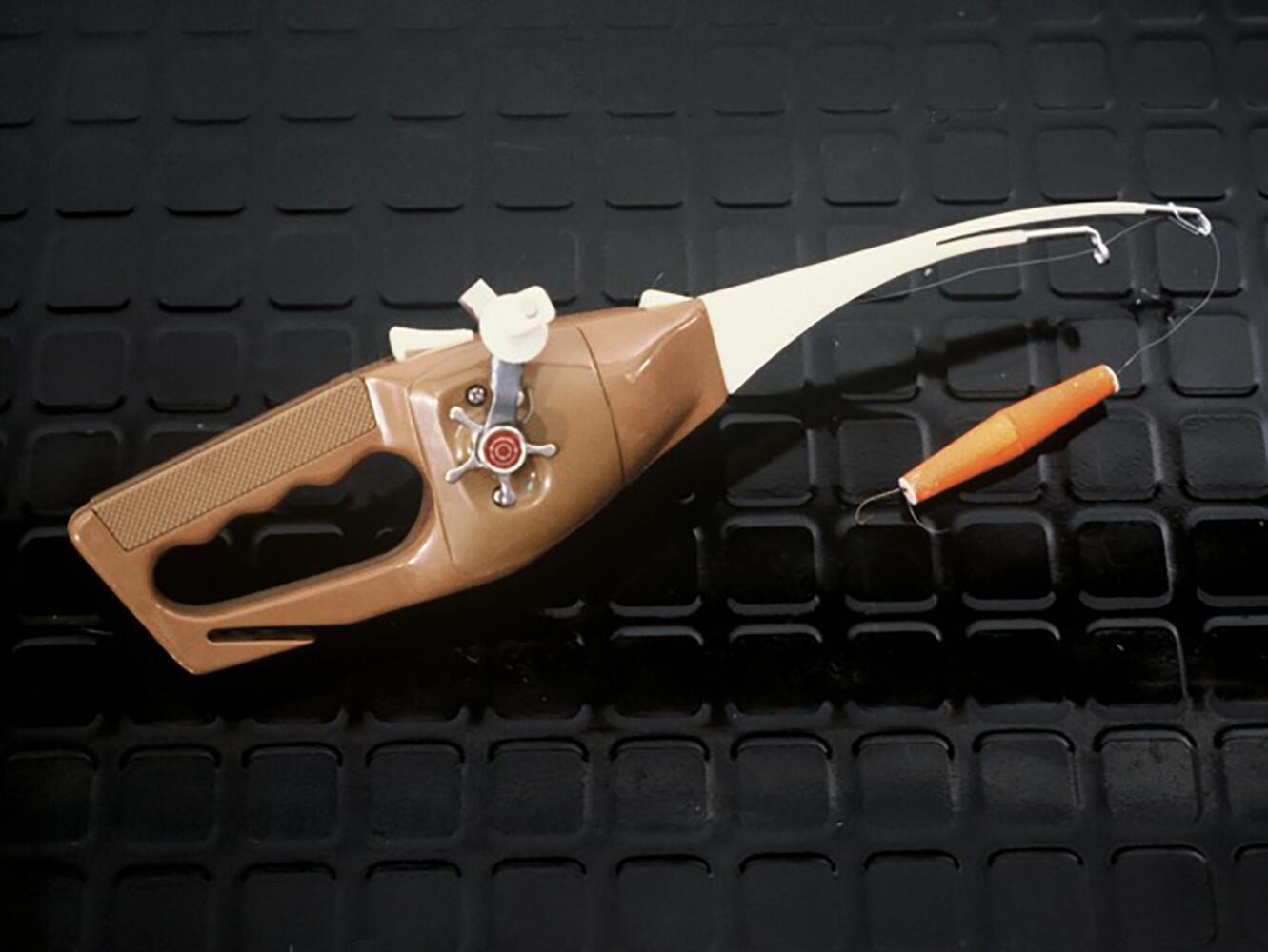 Brown fishing rod-style toy gun with a plastic handle, beige curved tip, and orange cork-like projectile on a string, placed on a textured black surface.