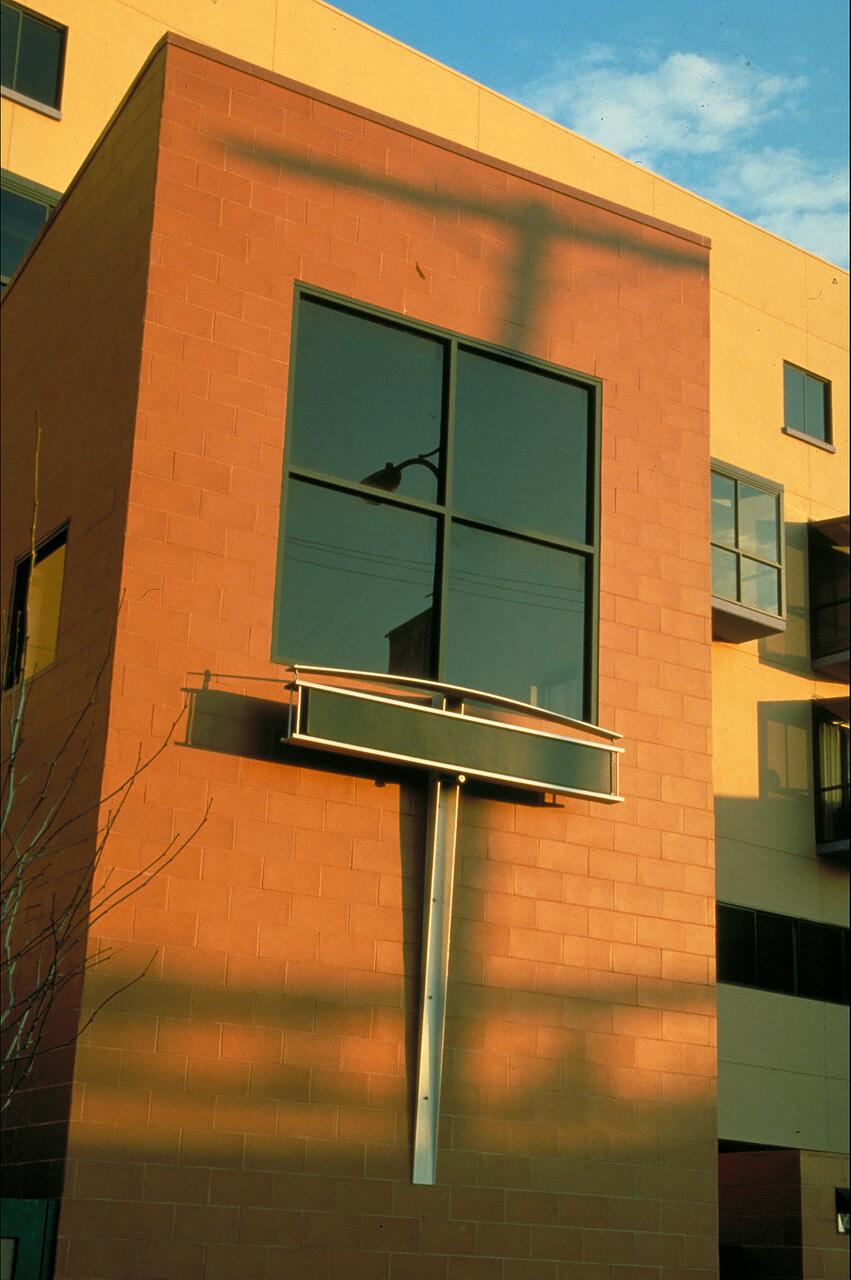 Modern building facade with a two-story salmon-colored wall, featuring large square windows. A protruding metal beam with a rectangular structure casts shadows, and the sky is clear with some clouds.