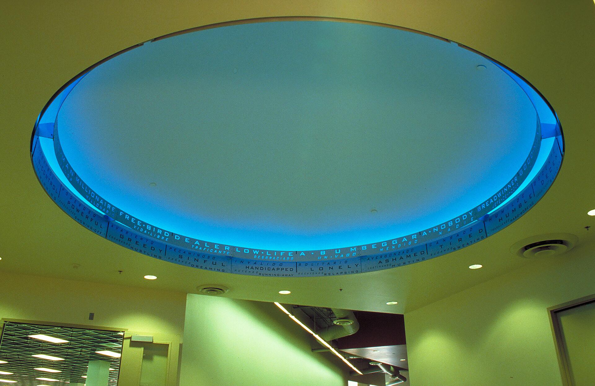 Circular ceiling light emitting a soft blue glow, surrounded by a band of words, in a room with beige walls and recessed ceiling lights.