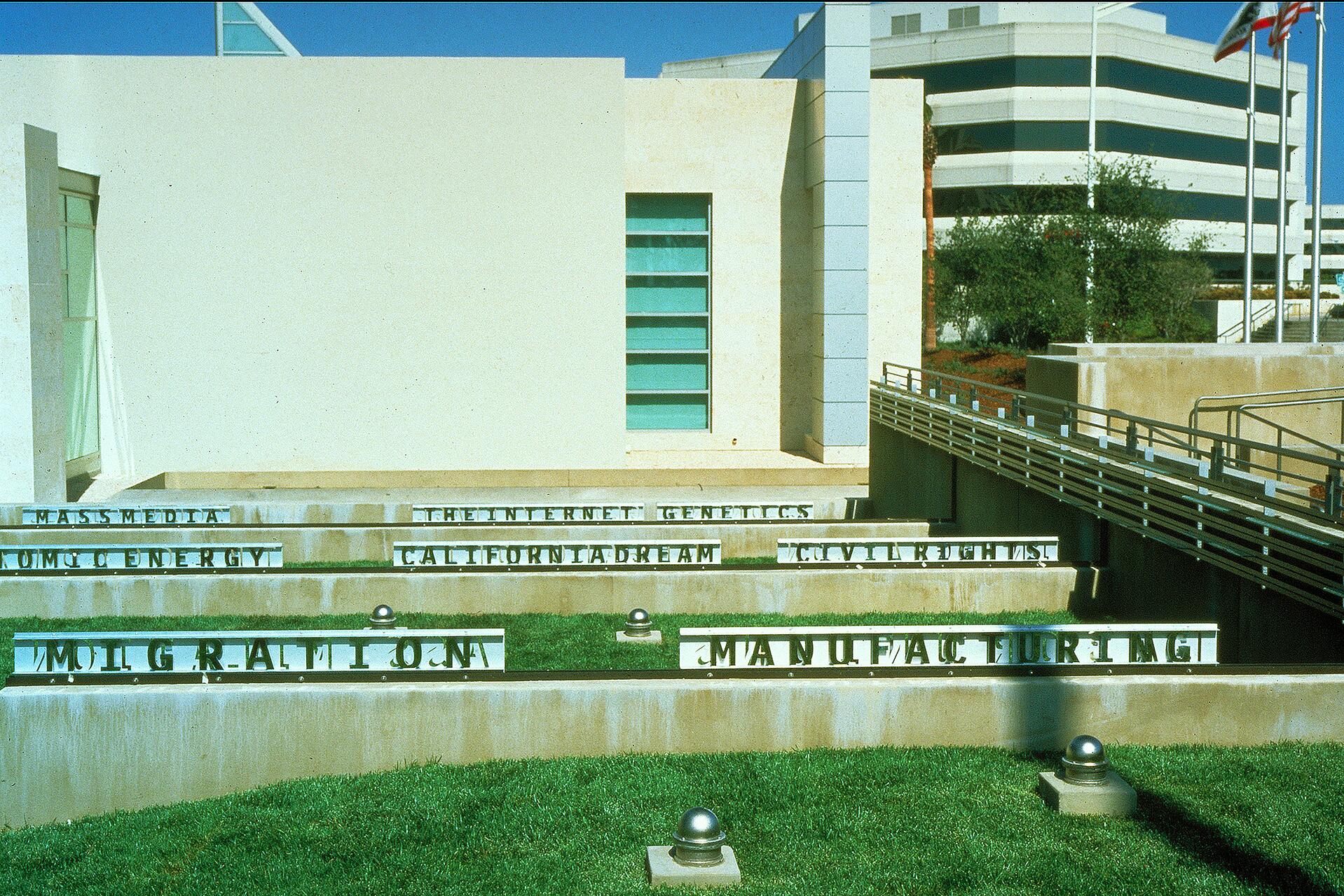 Modern building exterior with white walls and a green-tinted window. Concrete steps in the foreground display metal letters. Grass areas with small metal spheres interspersed. A multi-story office building and flagpoles are visible in the background.