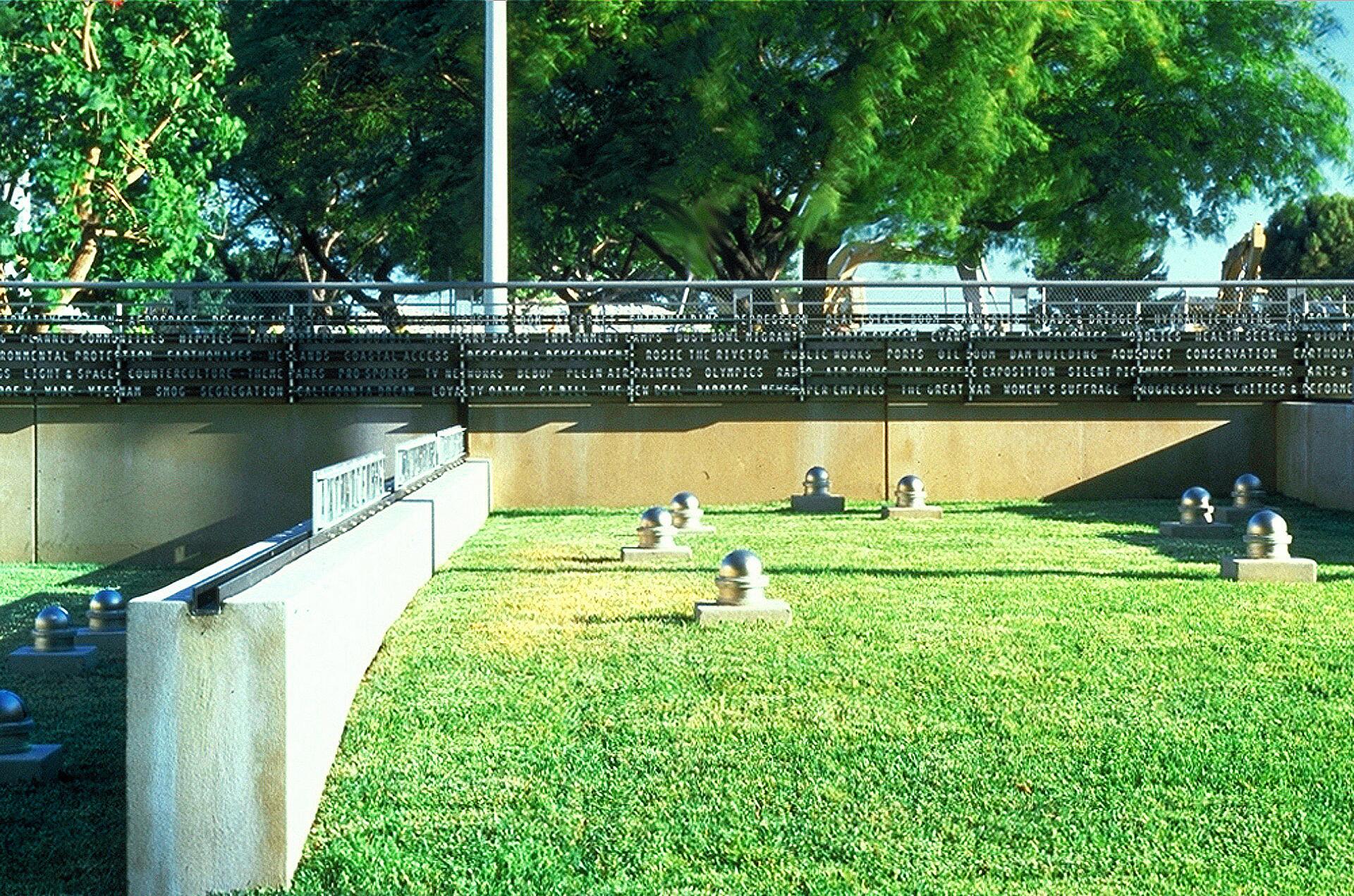Grassy courtyard with silver sphere sculptures on square pedestals, bordered by a low concrete wall. A black fence with cut-out letters creates shadows on the wall. Lush green trees are visible in the background.