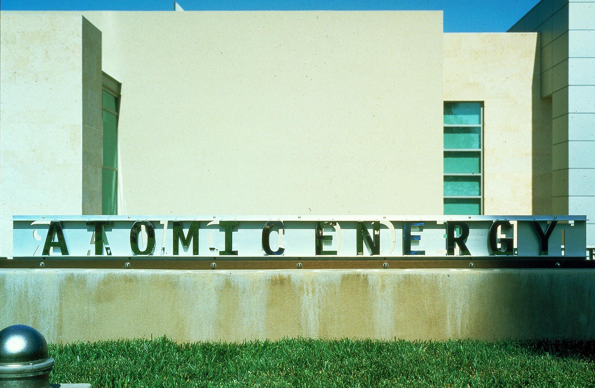 Sign with large letters spelling "ATOMIC ENERGY" on a grassy lawn, set against a modern beige building with blue sky above.