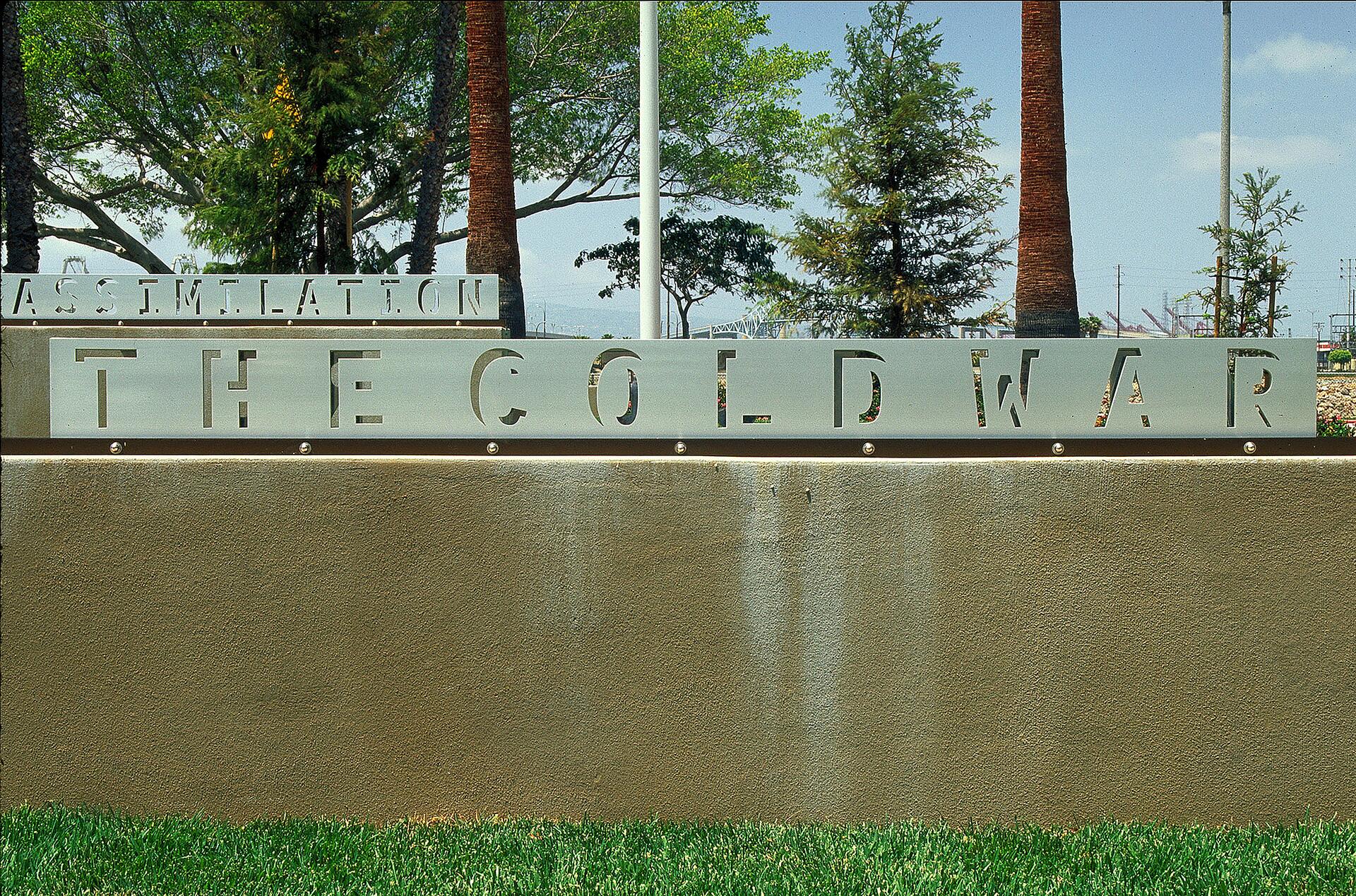 Gray metal sign with cut-out letters spelling "THE COLD WAR" against a concrete wall, set in a park with green grass and trees in the background.