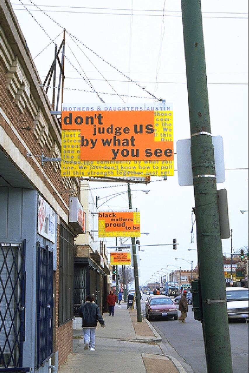 Street scene with pedestrians walking on a sidewalk. Bright, colorful signs with text hang above, attached to a brick building. Cars drive by on a road lined with various shops and businesses. A cloudy sky is visible above.