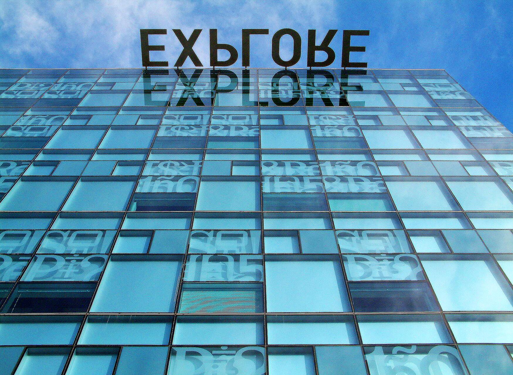 Glass building with "EXPLORE" written on top, reflecting on its surface. Geometric windows create a pattern, with blue sky background.