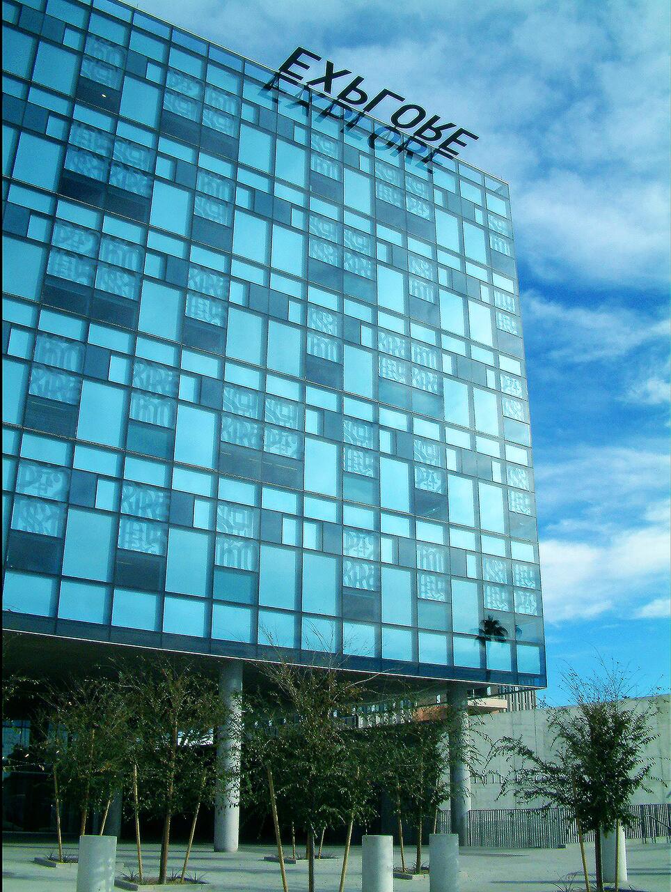 Modern glass building with a grid pattern reflecting the blue sky. Sparse trees line the foreground with a few clouds visible.