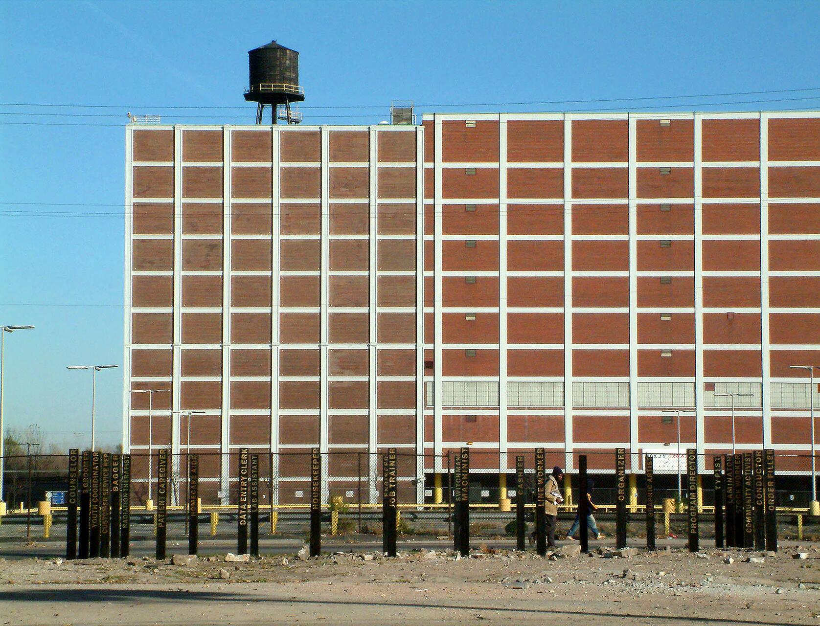 Brick building with a water tower on its roof against a clear blue sky. The foreground features a fenced area with multiple black poles, some with yellow text. Two people walk near the poles.