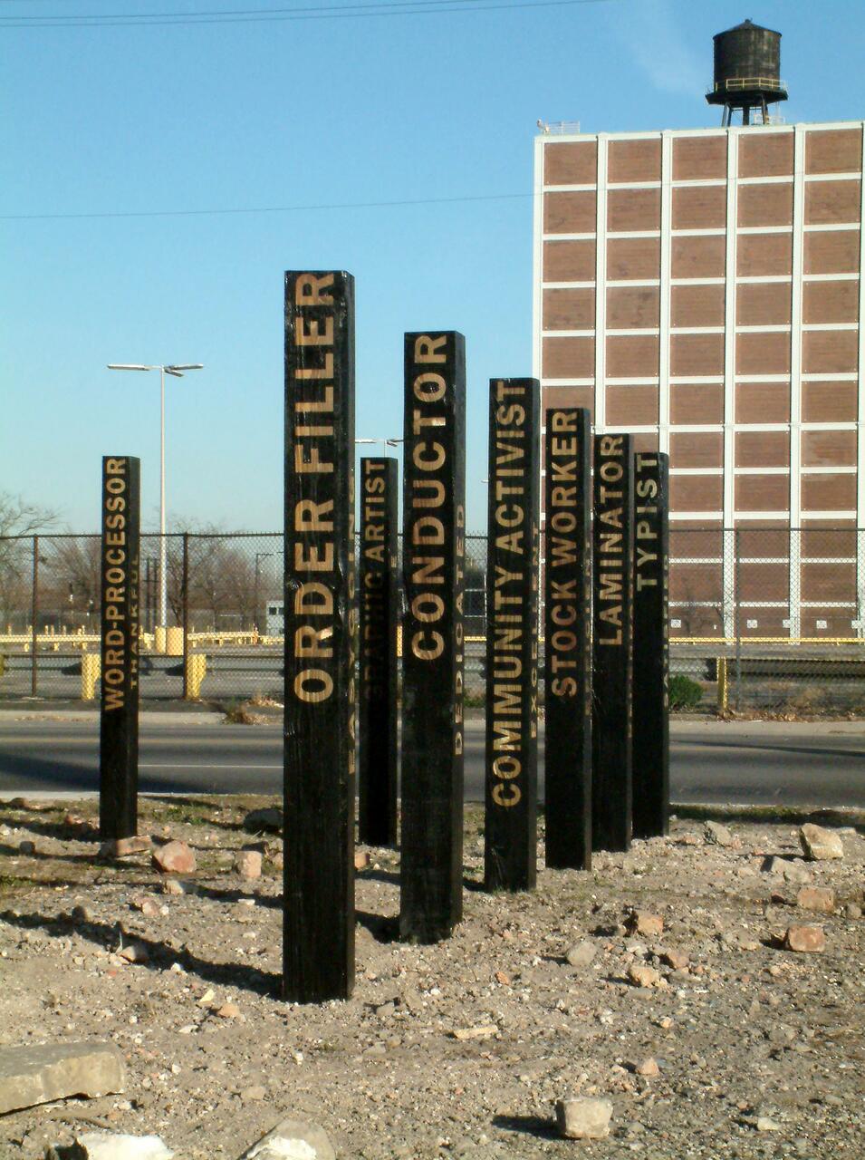 Black posts with text like "ORDER FILLER" stand upright in a barren lot. A large building and water tower are visible in the background under a clear blue sky.