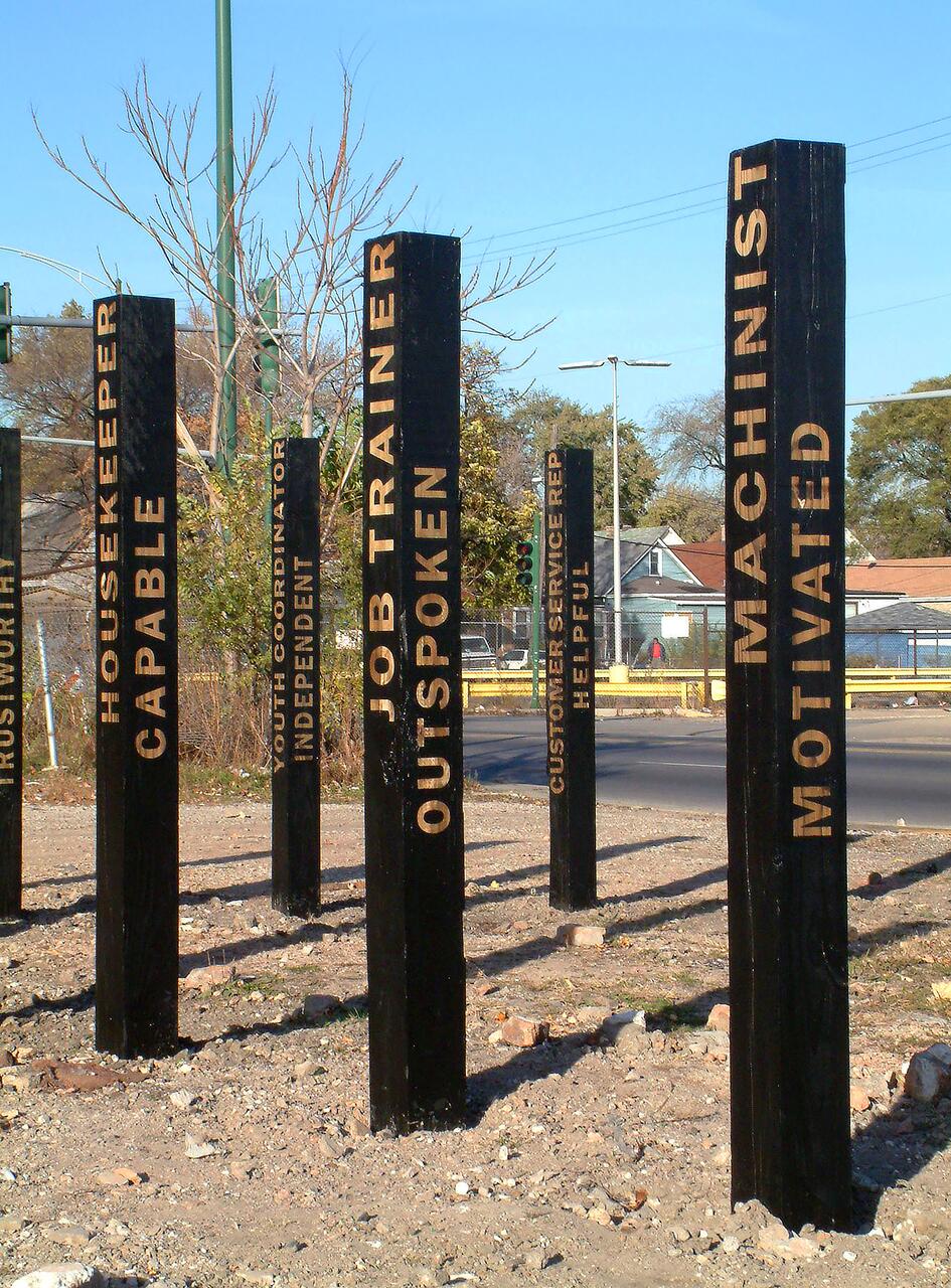 Tall black pillars in an outdoor setting, each inscribed with words like "Job Trainer," "Machinist," and "Housekeeper." The sky is clear and blue, and some trees and buildings are visible in the background.