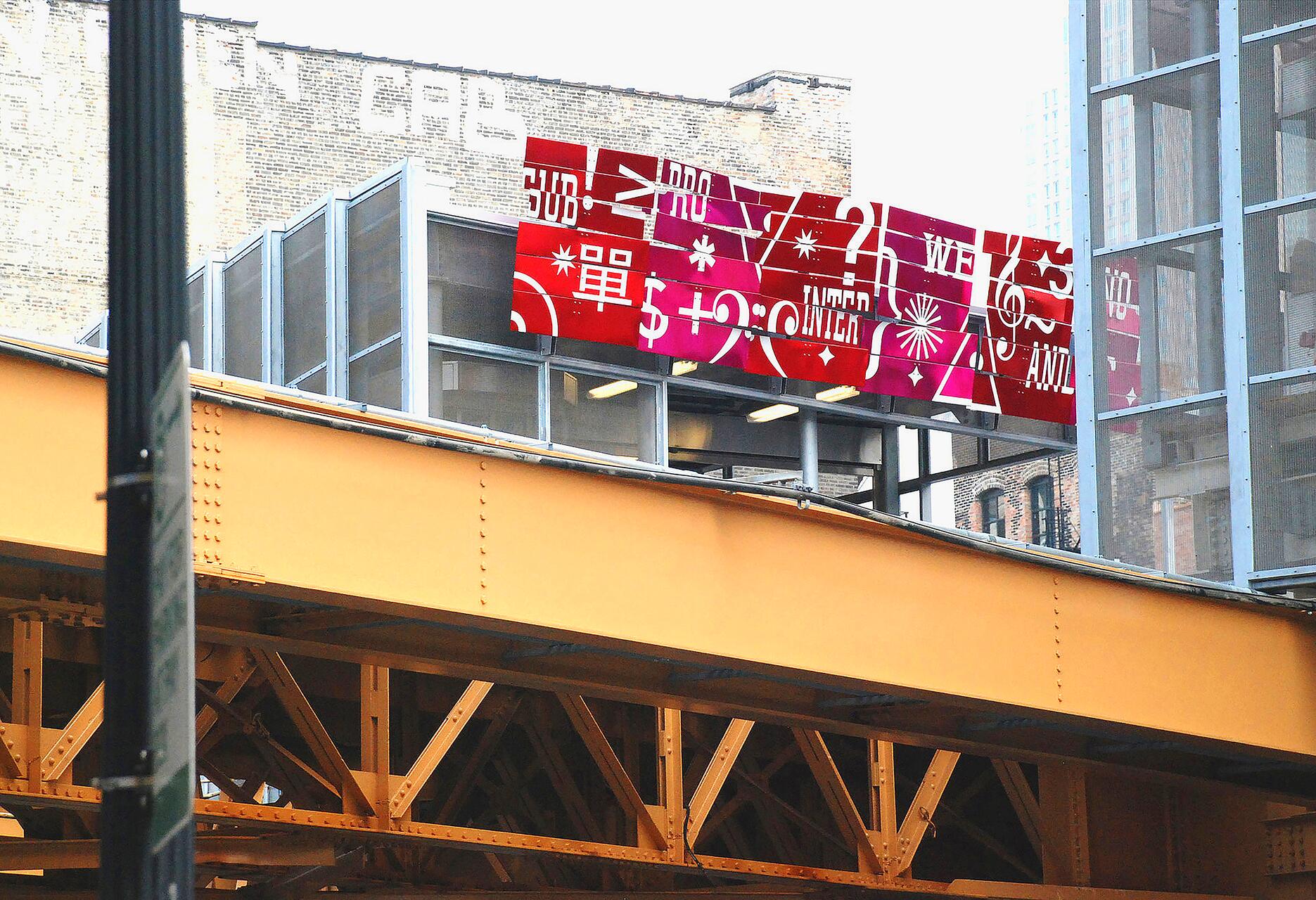 Urban scene with a modern red and white graphic sign featuring symbols and numbers above a yellow elevated railway track. A brick building and glass structure are in the background.