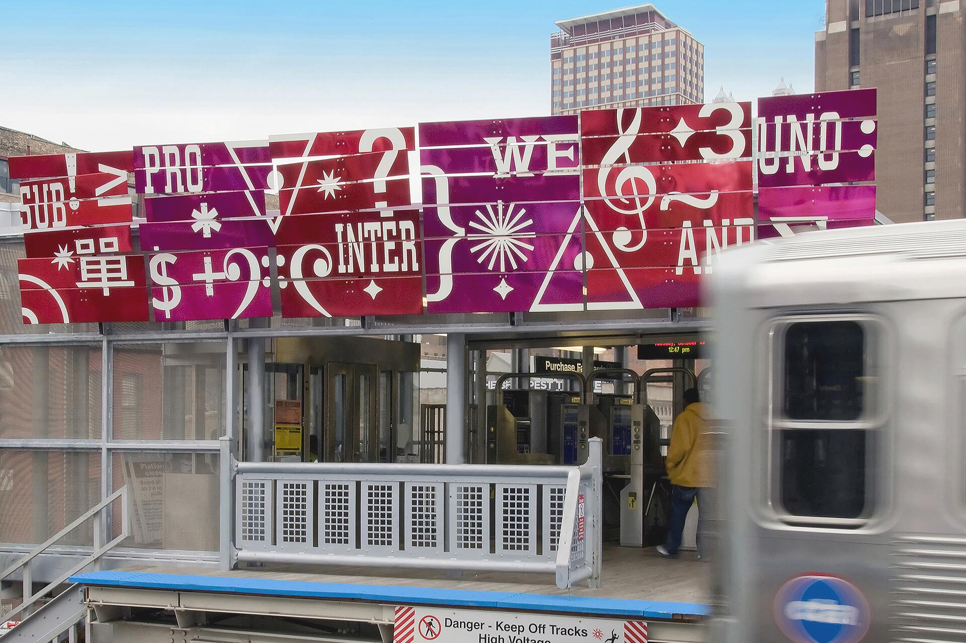 Colorful mural with various symbols and words above a train station entrance, a train passing by in the foreground. A person in a yellow jacket is entering through the turnstile. Tall buildings in the background.
